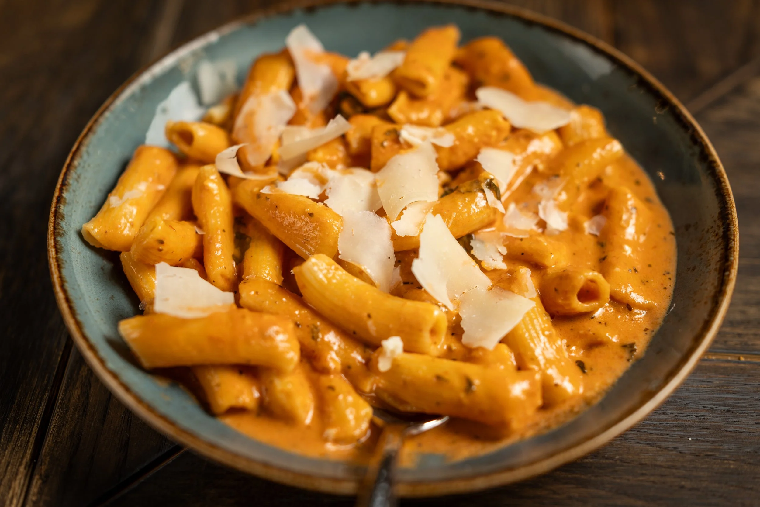 A bowl of pasta with orange-colored creamy tomato sauce topped with shaved cheese, on a dark wooden surface.