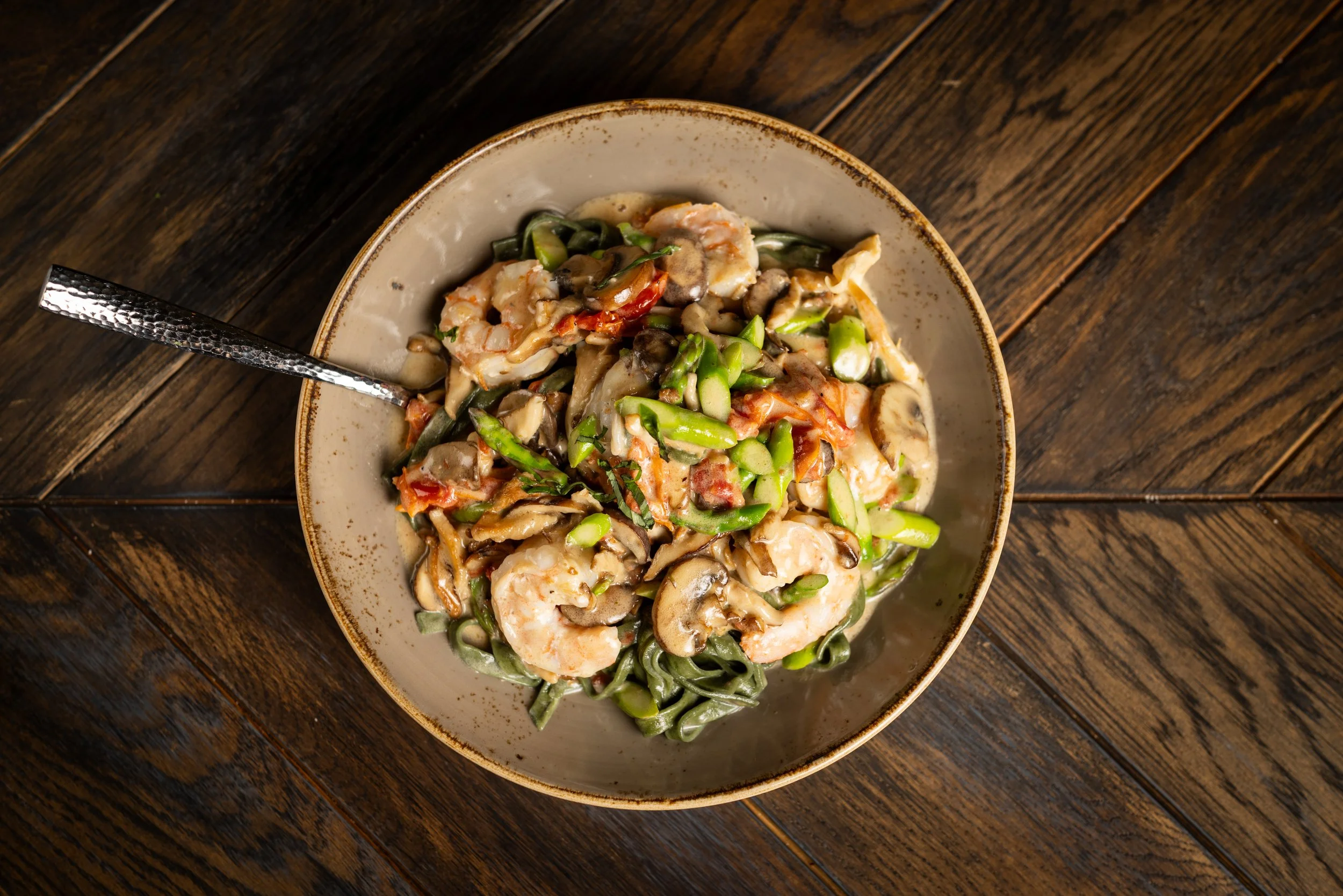A bowl of pasta with shrimp, mushrooms, green beans, and a creamy sauce garnished with herbs on a wooden table.