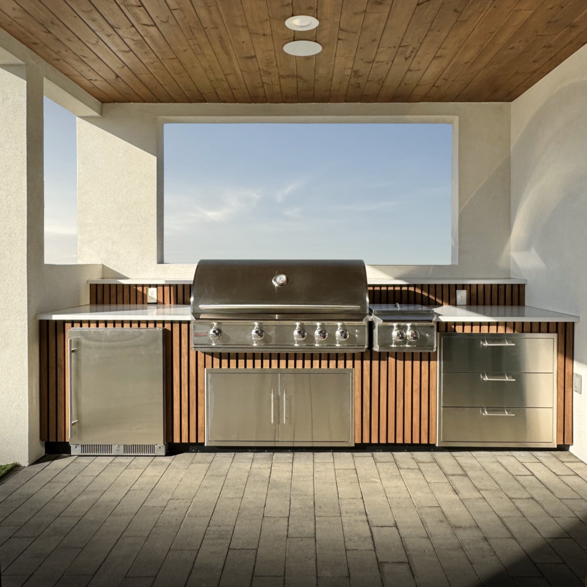 Outdoor kitchen with stainless steel grill and wooden paneling beneath a wooden ceiling, open to the sky.