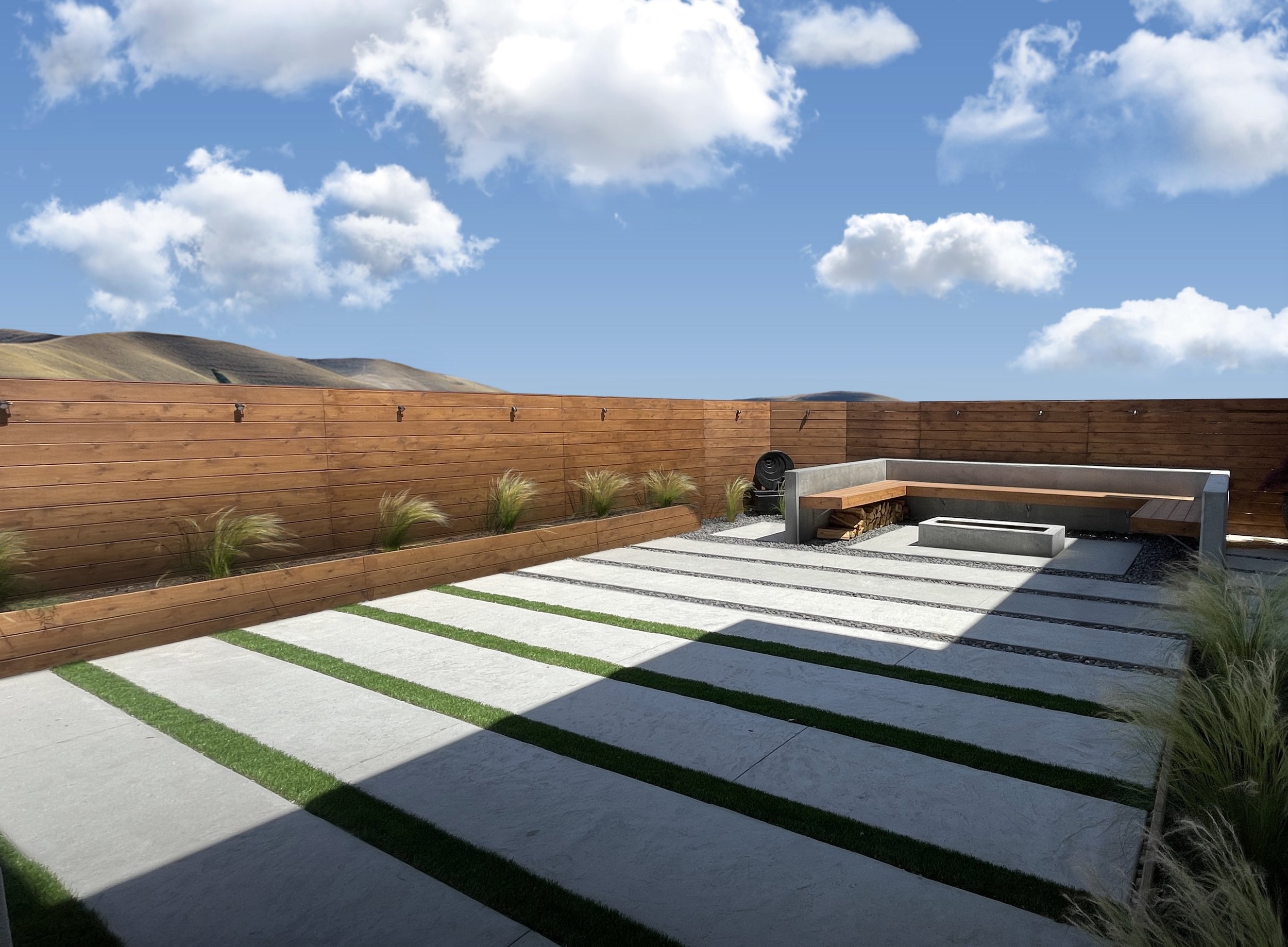 A backyard with a tall wooden privacy fence, geometric concrete and grass patio, and a seating area with benches. There are small plants along the fence and rolling hills in the background under a blue sky with white clouds.