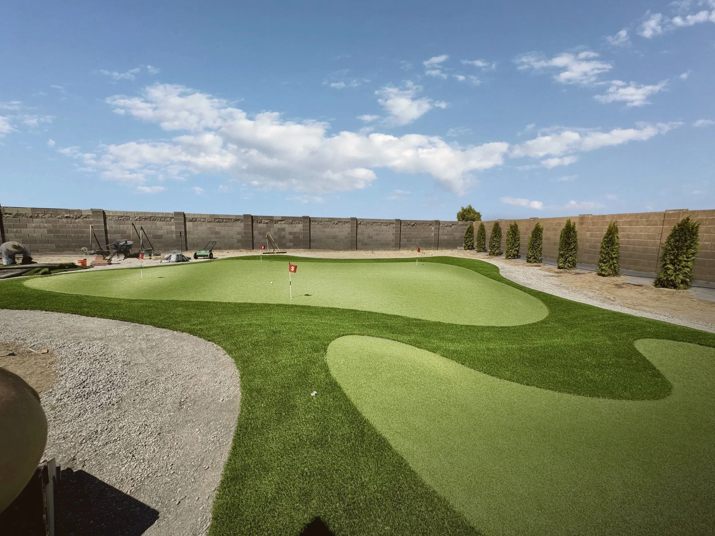 A golf course putting green with flags, surrounded by a gravel pathway, a row of tall, evenly spaced trees along a brick wall, and a clear blue sky with scattered clouds.