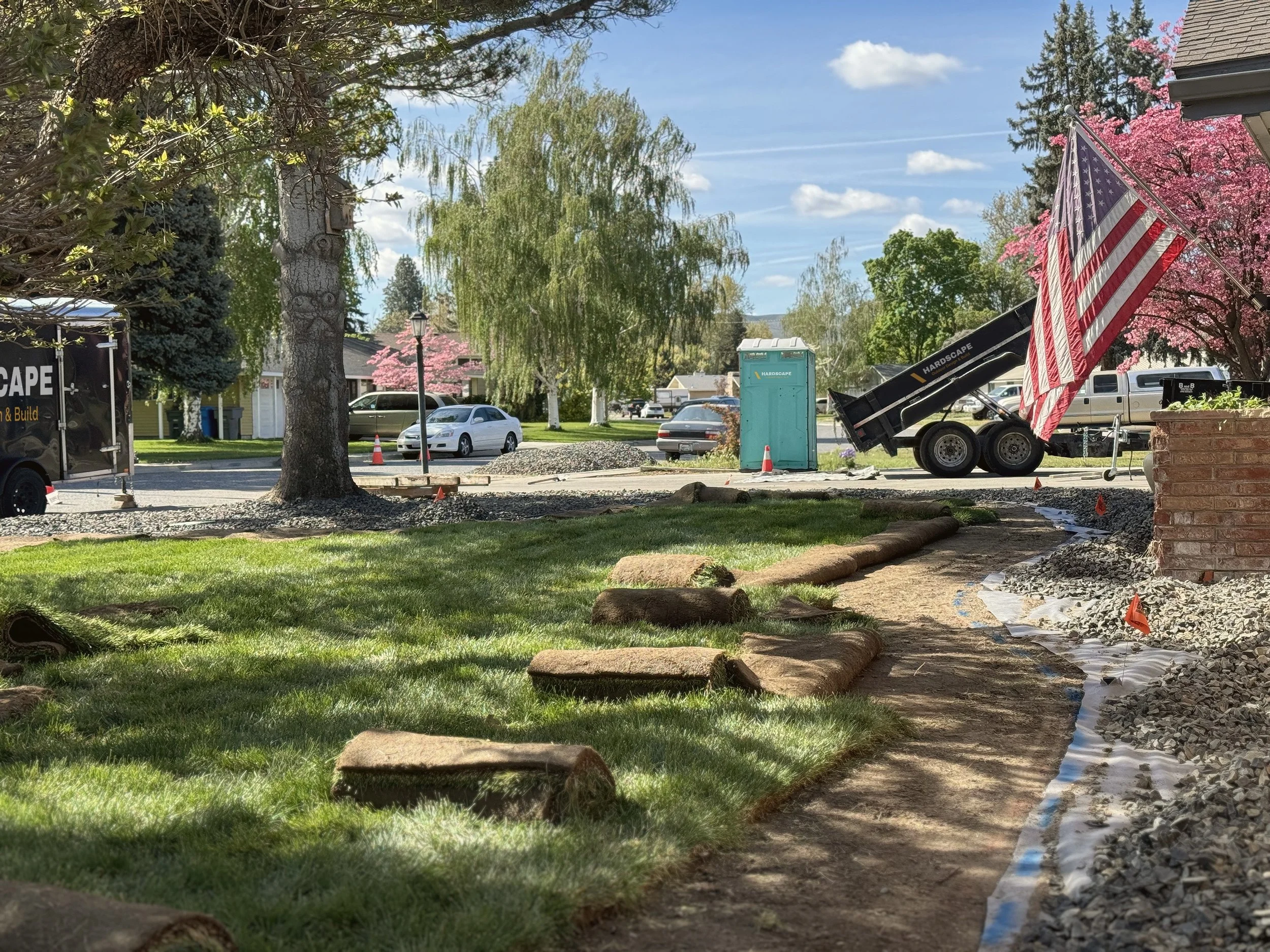 A residential street under renovation with work equipment, a portable toilet, white and gray parked cars, trees with pink flowers, and a blue sky with white clouds.