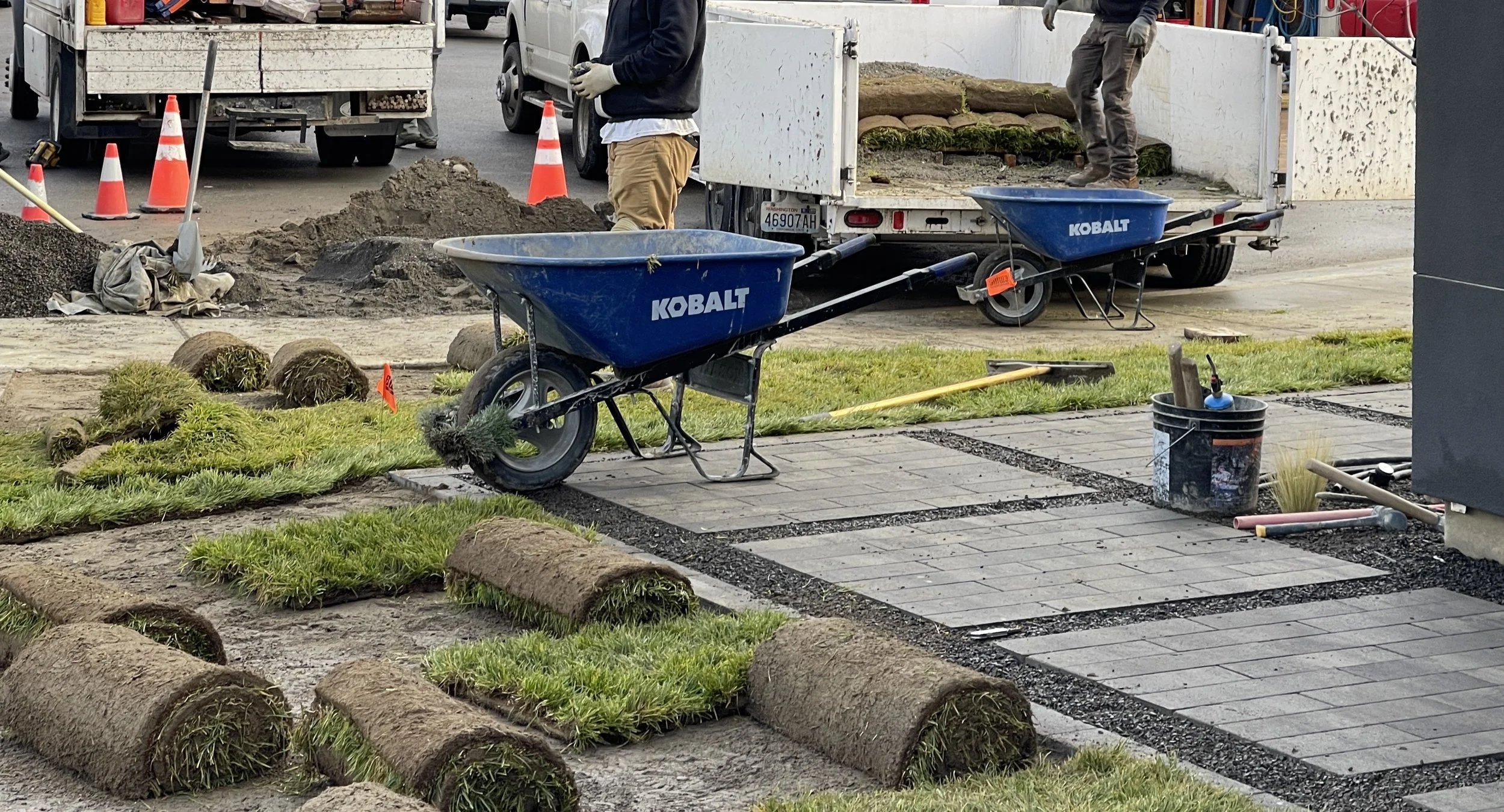 Workers installing pavers and landscaping a lawn area, with tools and wheelbarrows nearby, in an urban setting.