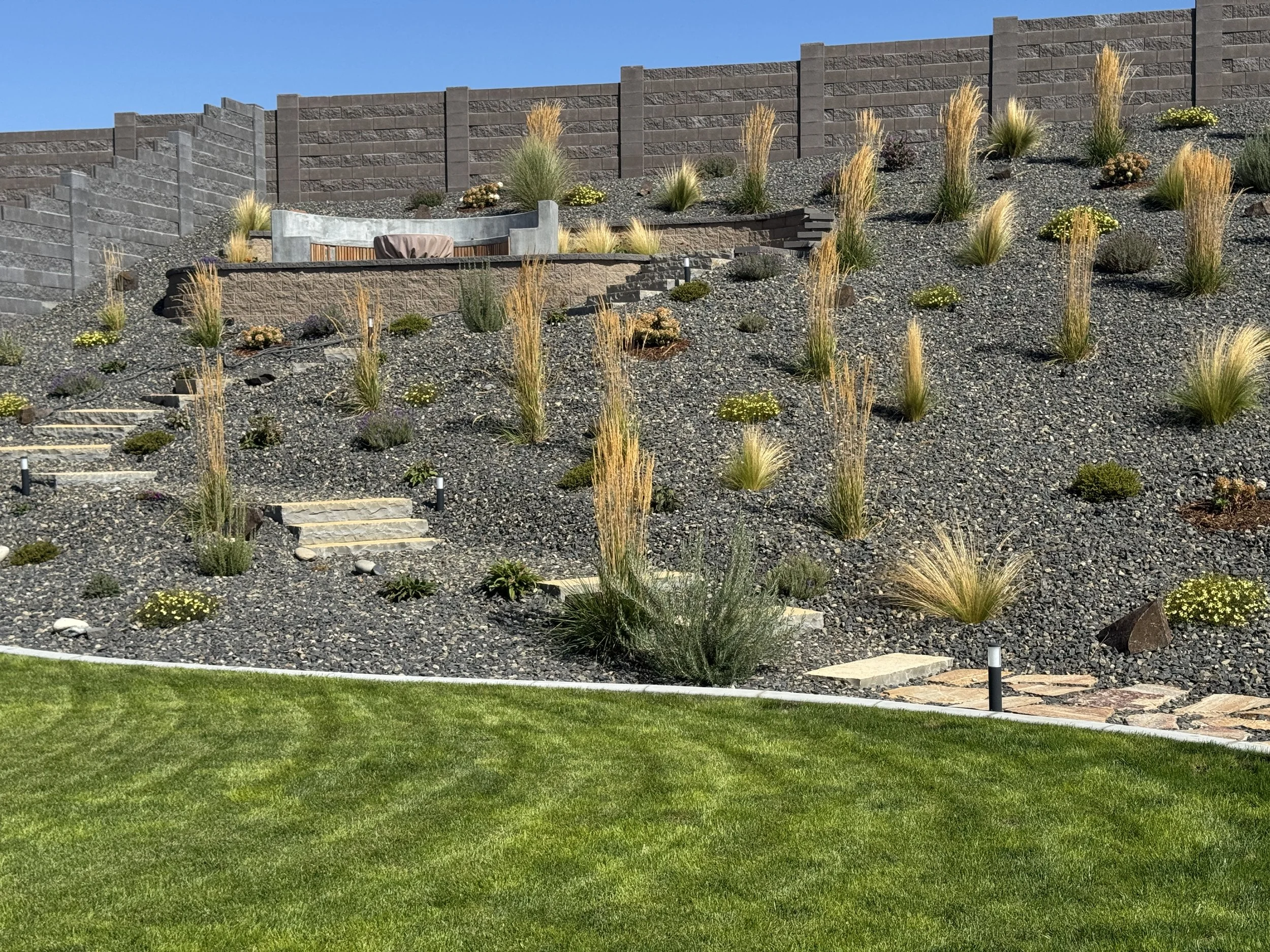A landscaped backyard with a tiered stone retaining wall, desert plants, and outdoor seating area against a blue sky.