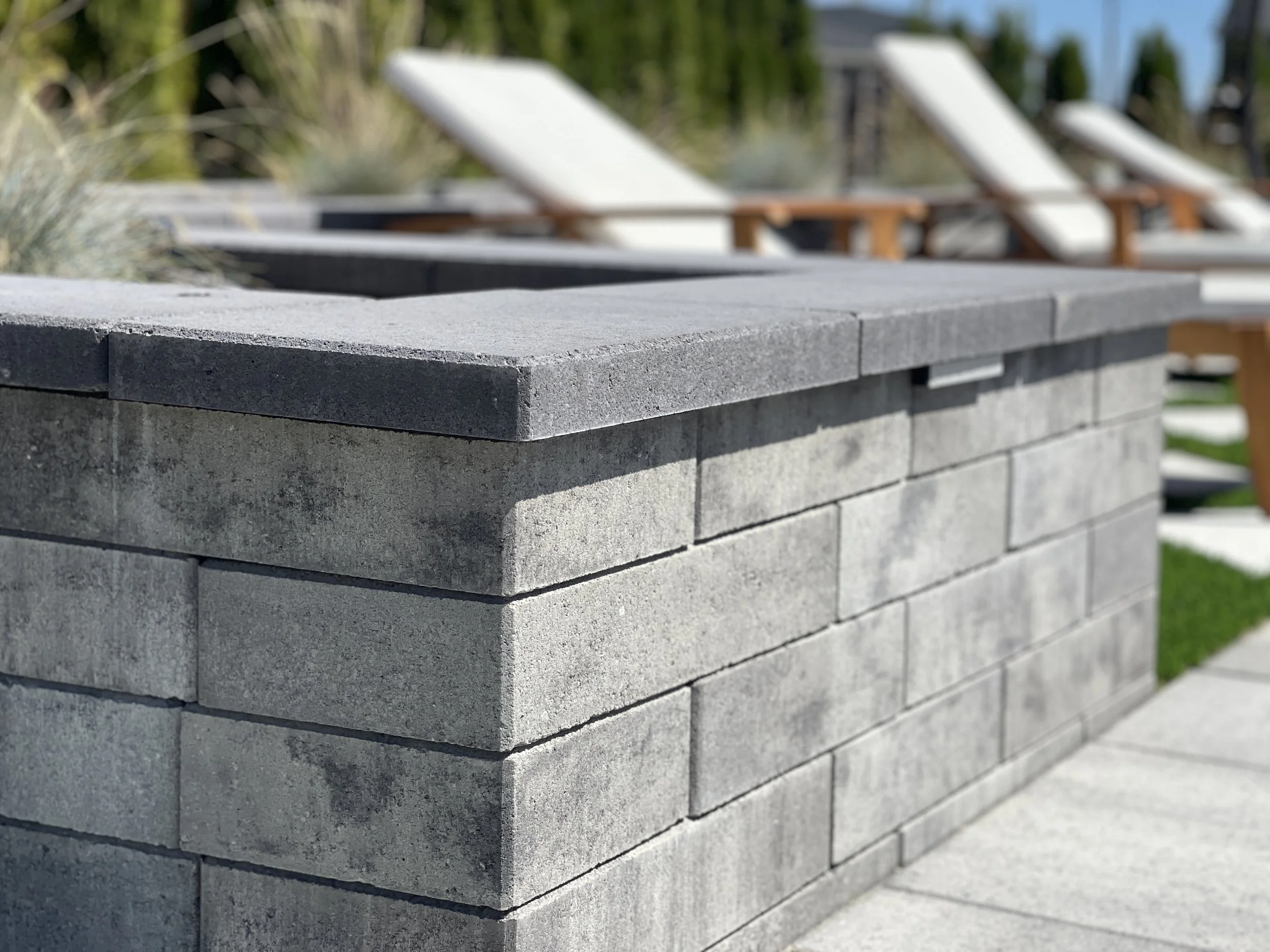 A close-up of a gray brick wall with several bright white lounge chairs in the background, outdoors on a sunny day.