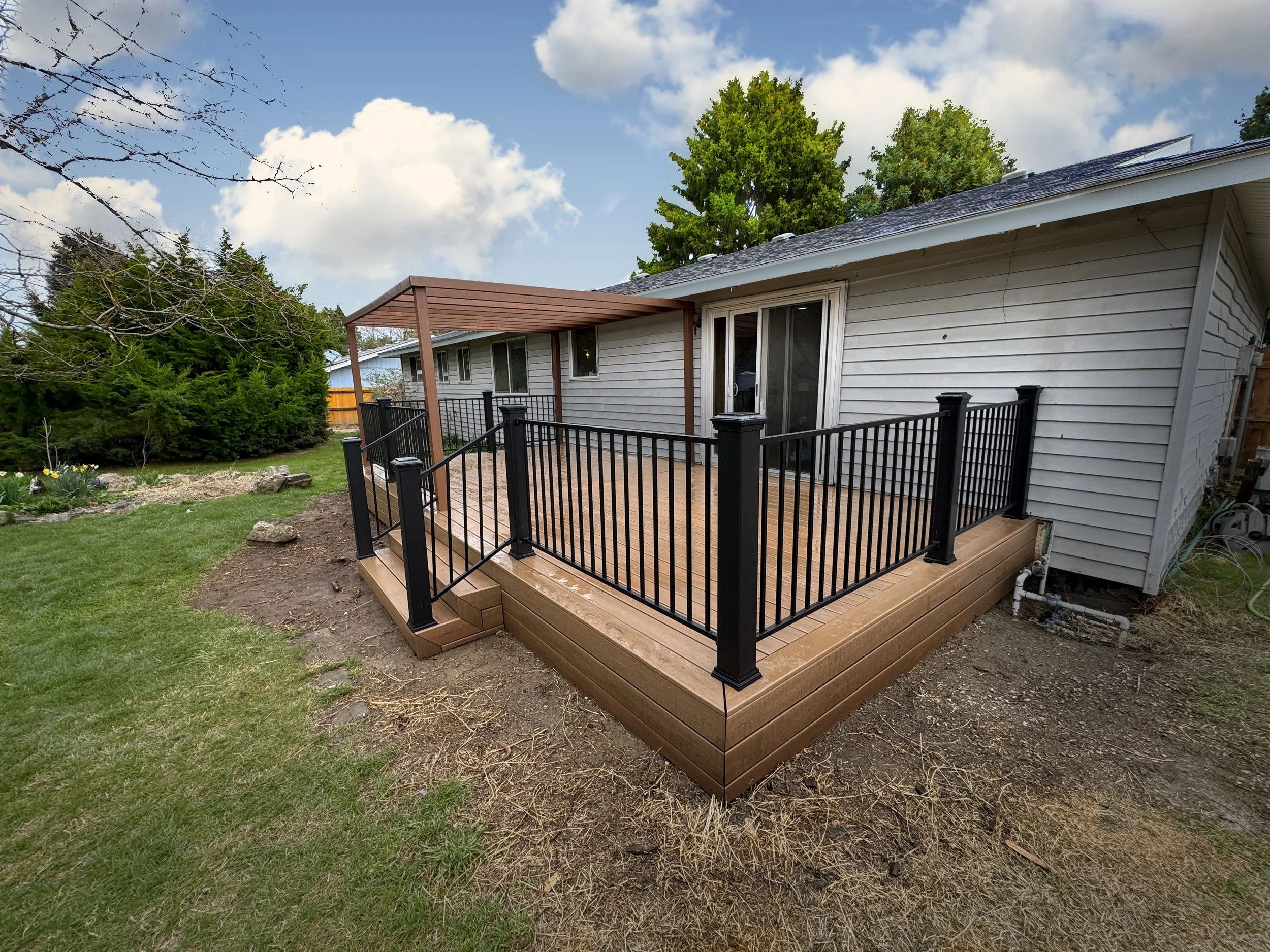 A backyard patio with a new wooden deck, black metal railing, and a wooden pergola. The house has white siding, sliding glass door, and a roof with skylights. Green trees, shrubs, and cloudy sky are in the background.