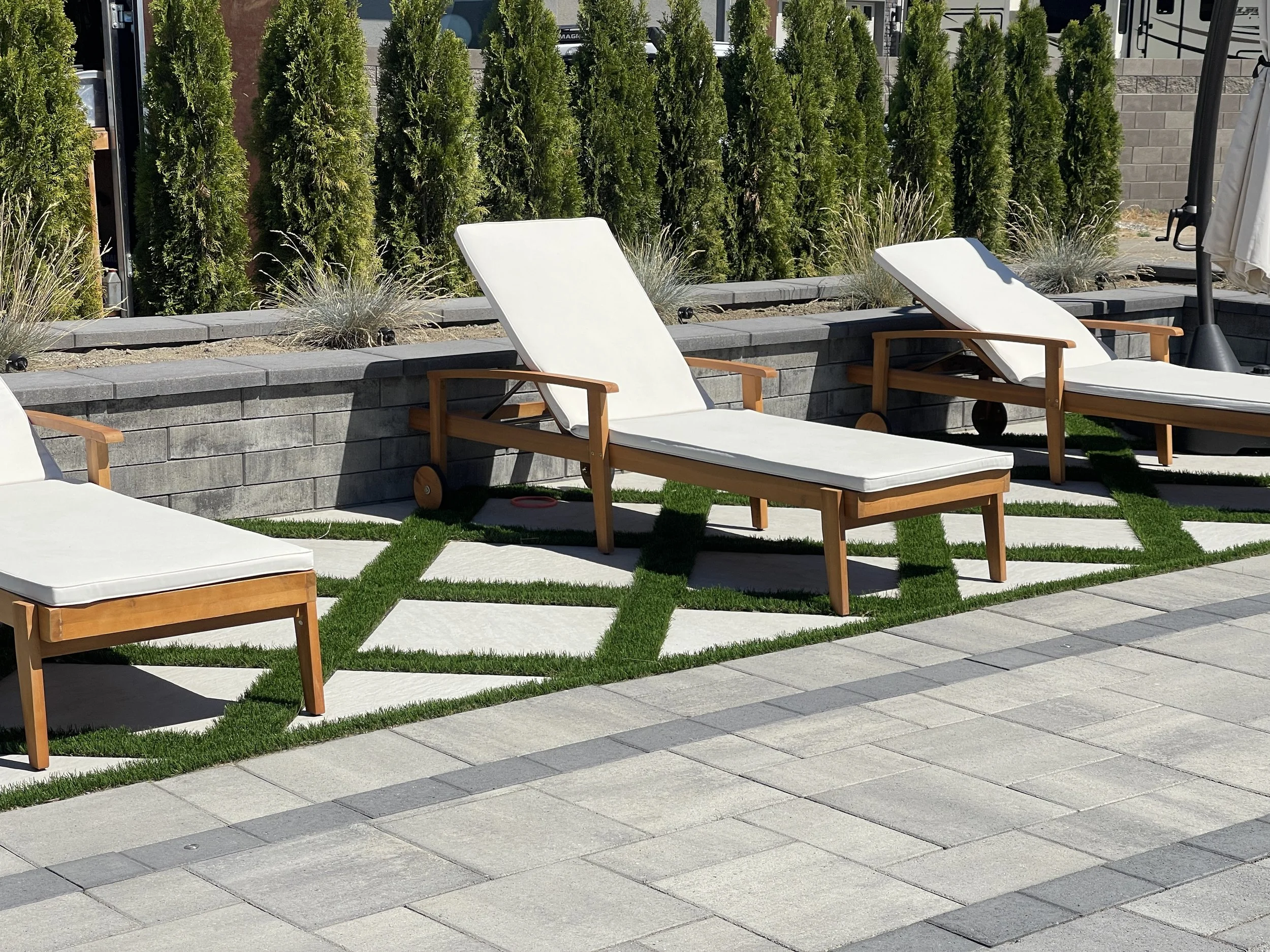 Outdoor patio with two white lounge chairs with wooden frames placed on a paved surface, enclosed by a low gray stone wall with tall green shrubs behind it, and potted ornamental grass plants. There is a barbecue grill and patio umbrella on the right