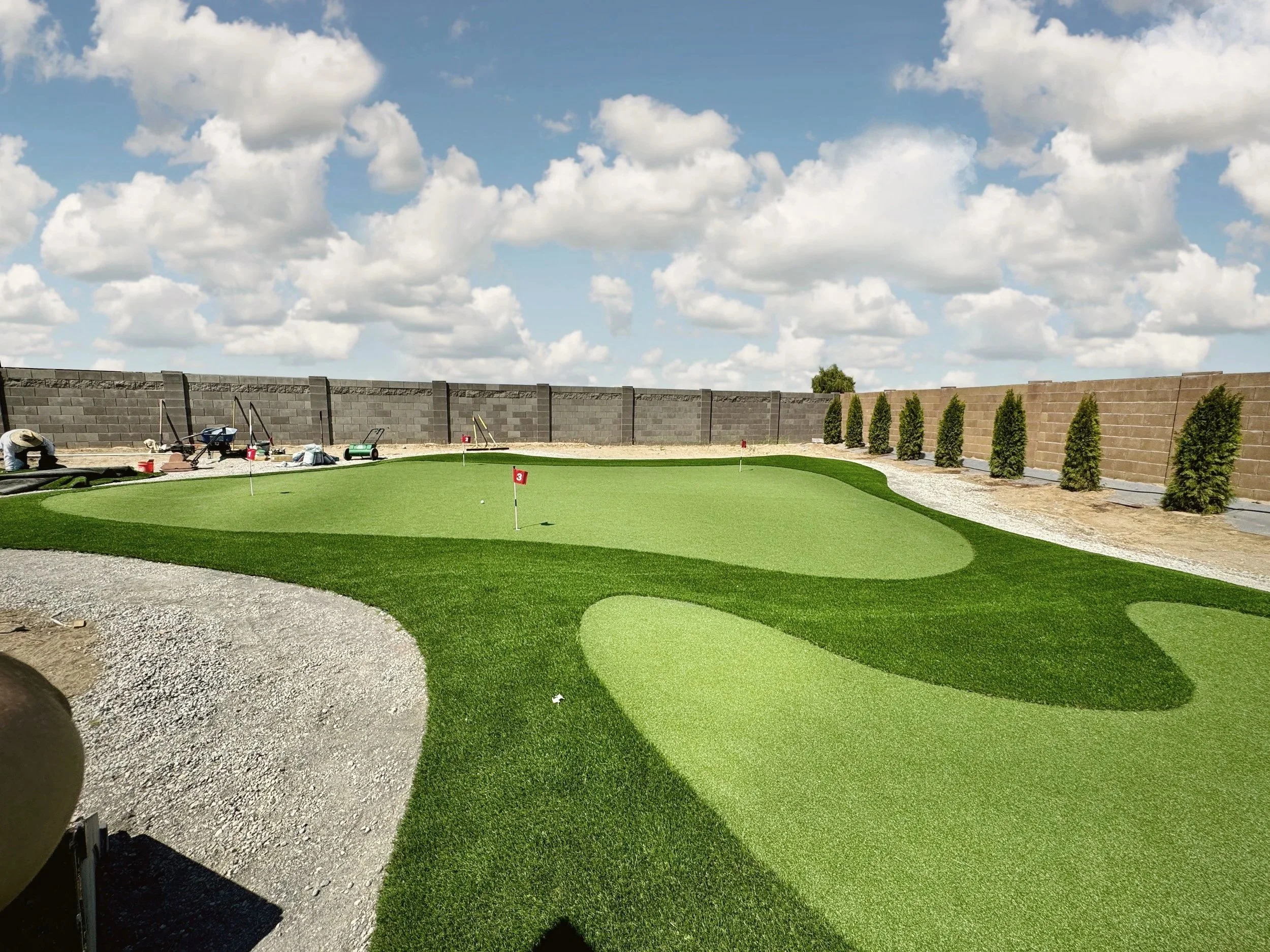 A golf course putting green with flags marked 3, surrounded by a gravel path, and a line of small trees against a cloudy sky.