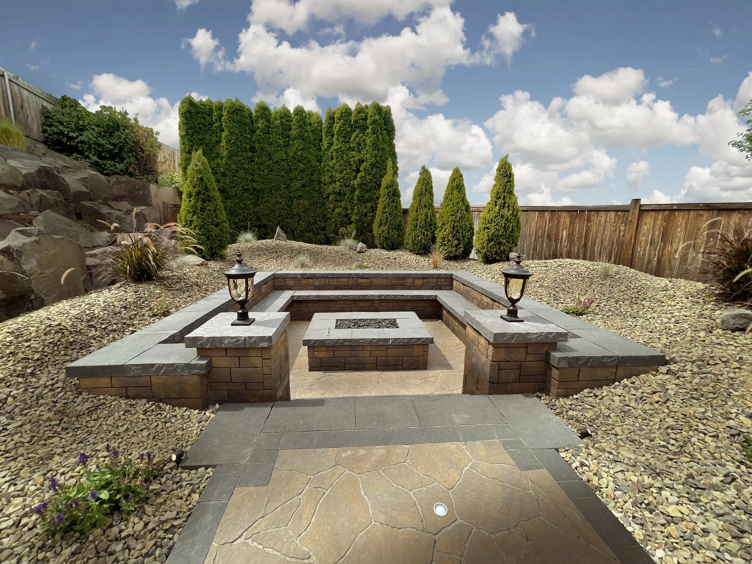 Backyard with stone steps leading to a patio, surrounded by rocks, small plants, and tall green trees, with a wooden fence and partly cloudy sky overhead.