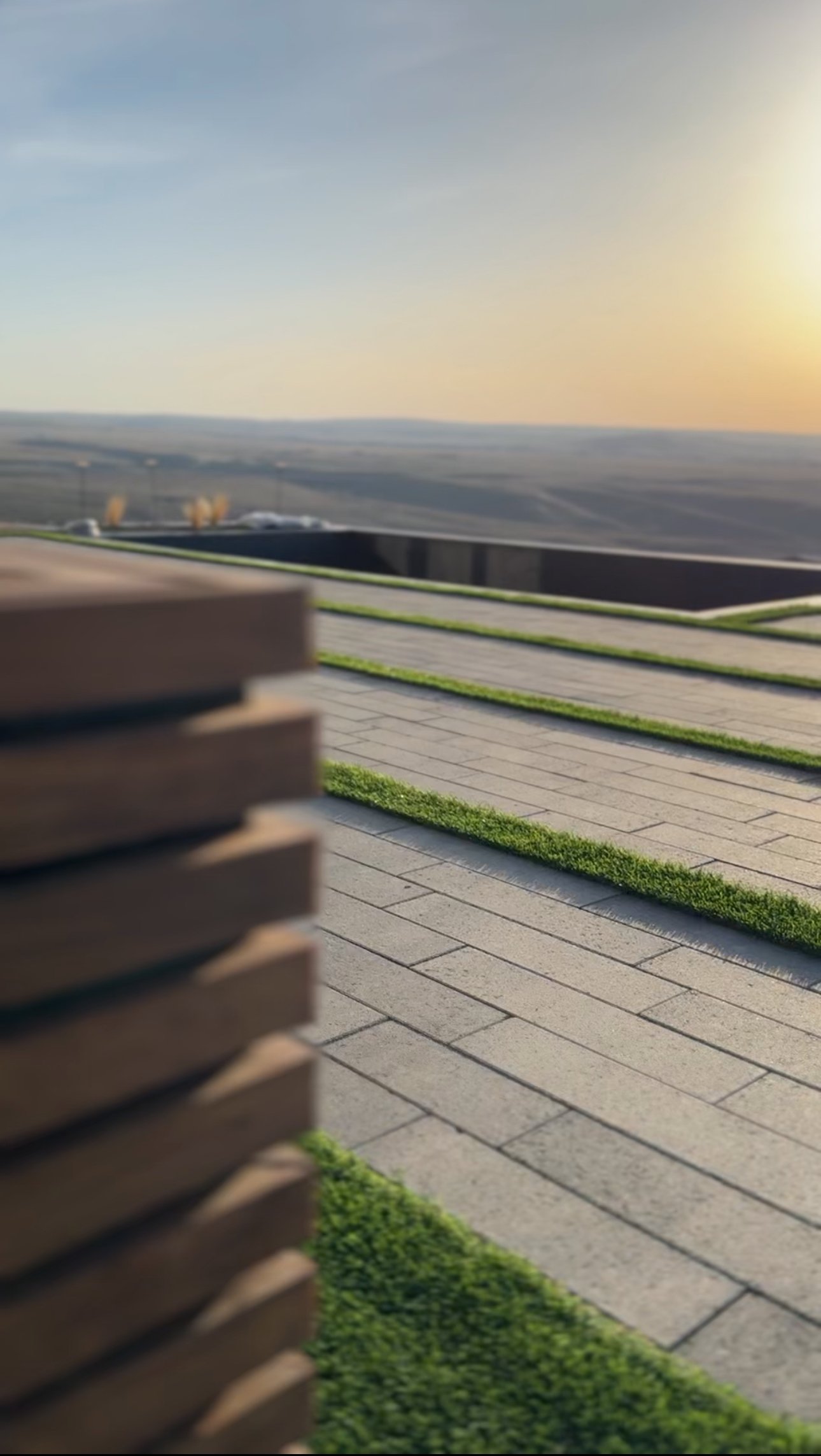Outdoor scene with a wooden bench and paved walkway, green grass, and a distant landscape under a clear sky at sunset.