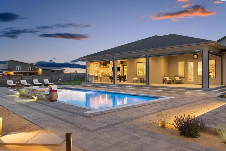 Backyard with a lit swimming pool, outdoor lounge chairs, and a house with large windows and a patio at dusk.