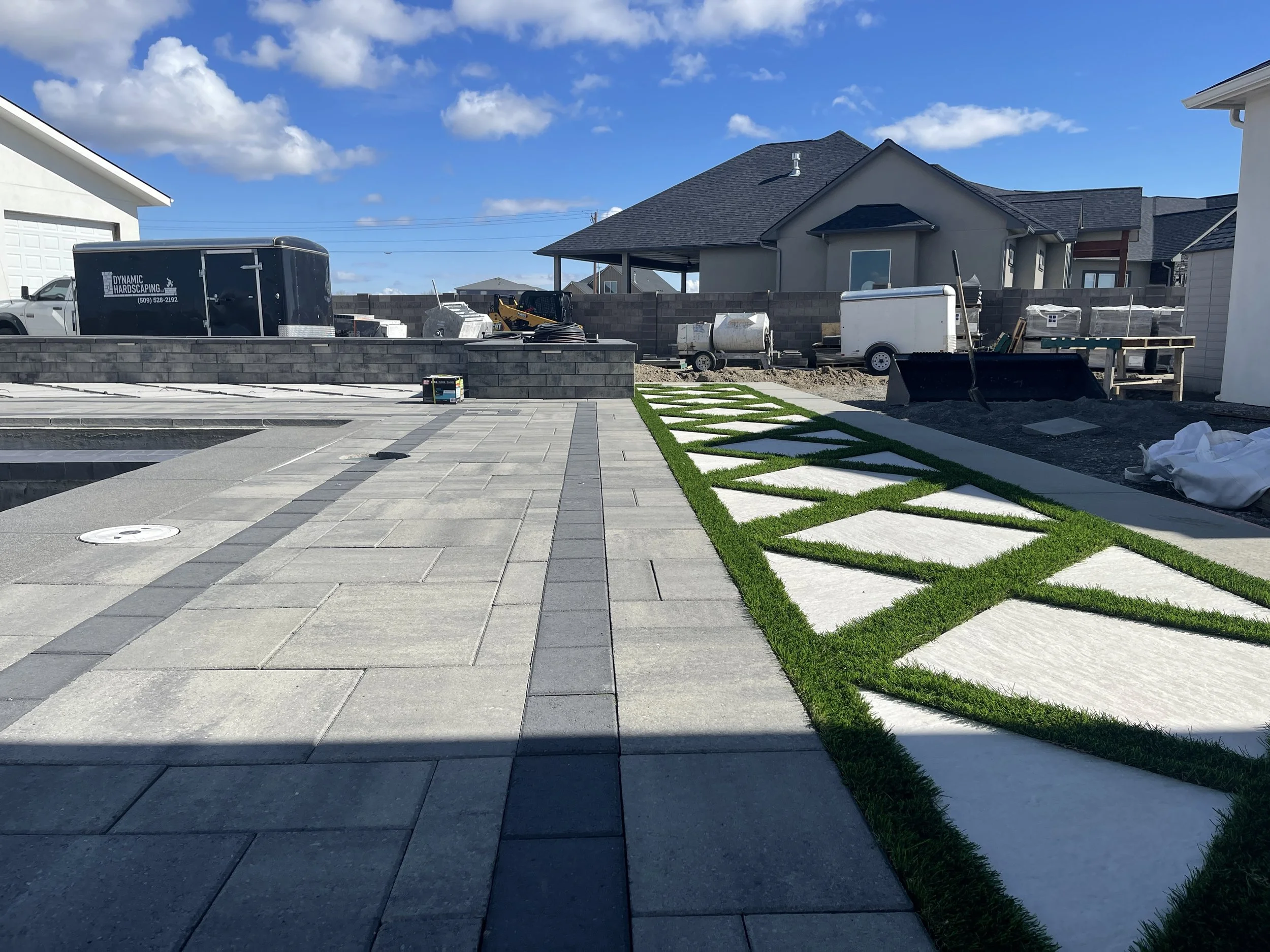 A backyard under construction with paving stones, green grass, construction equipment, a trailer, and a partially finished brick wall, with houses and a blue sky with clouds in the background.