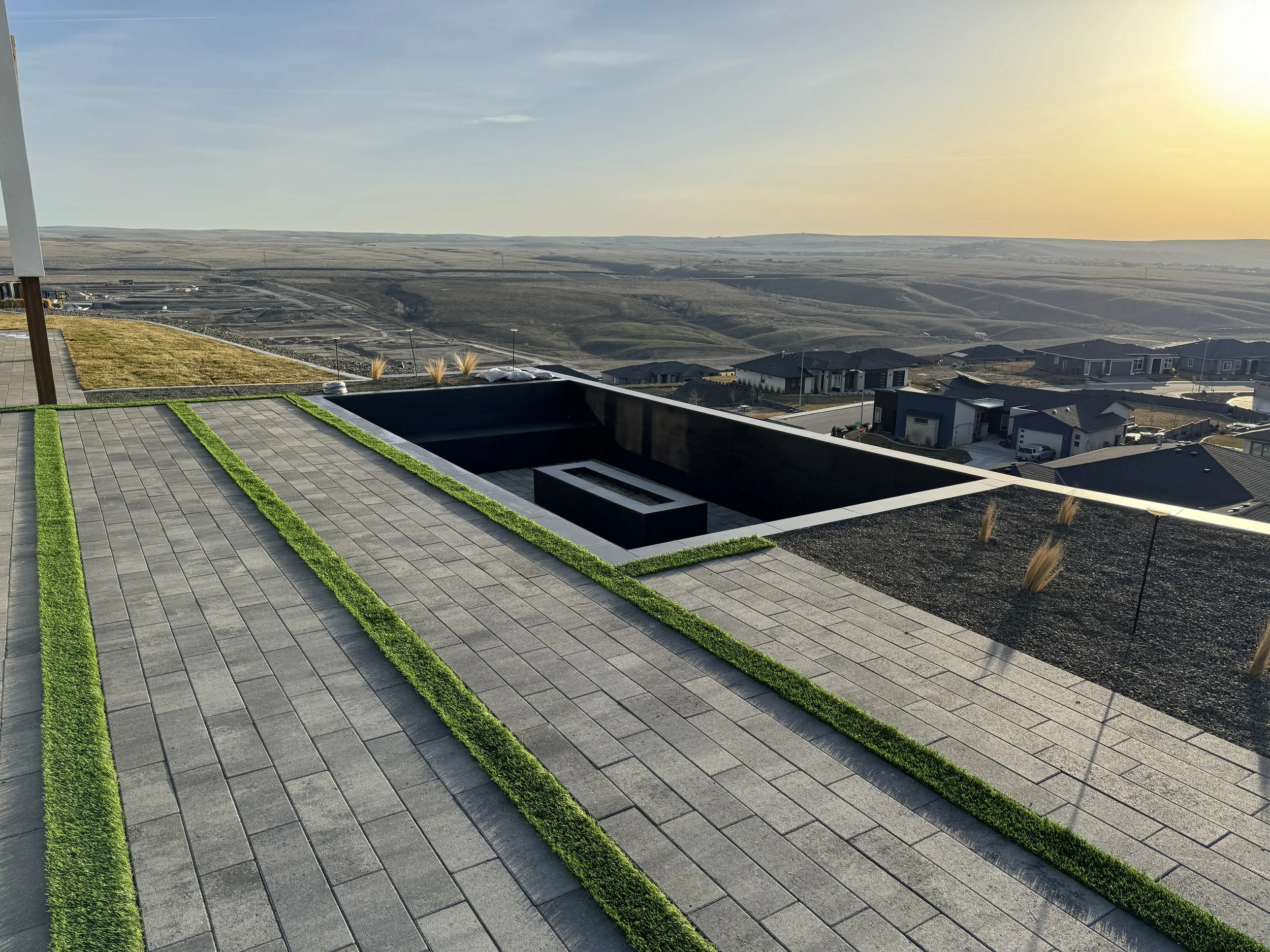 A modern house rooftop with paved tiles and strips of green grass, overlooking a suburban neighborhood with rolling hills and a setting sun in the distance.