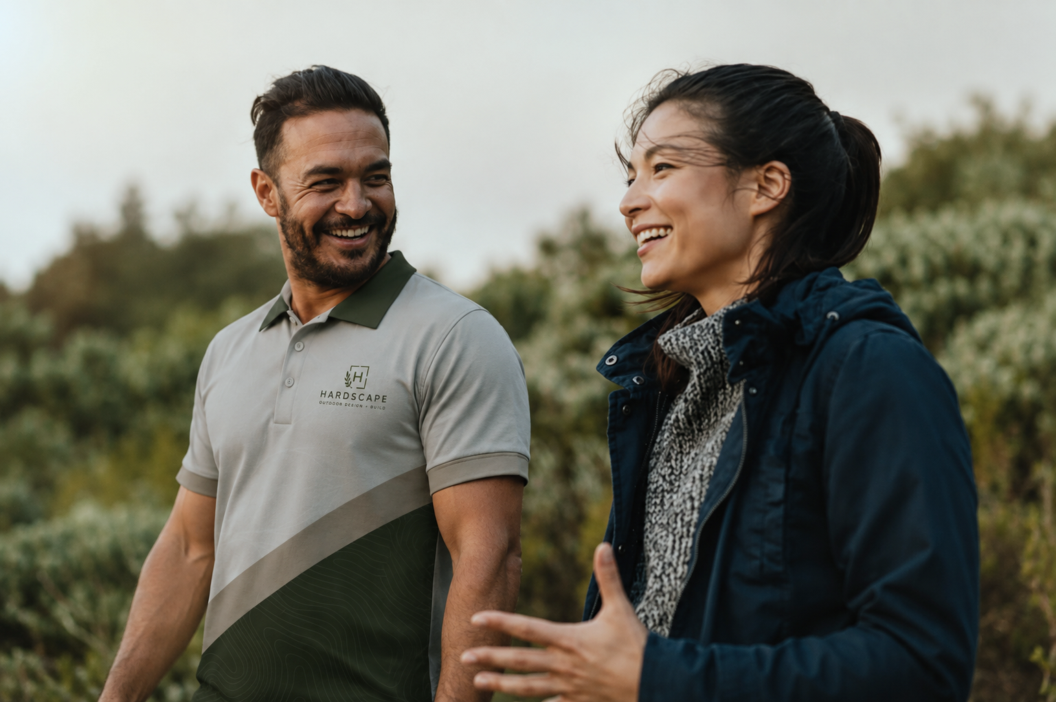 A man and woman smiling and walking outdoors in a natural setting with greenery in the background.