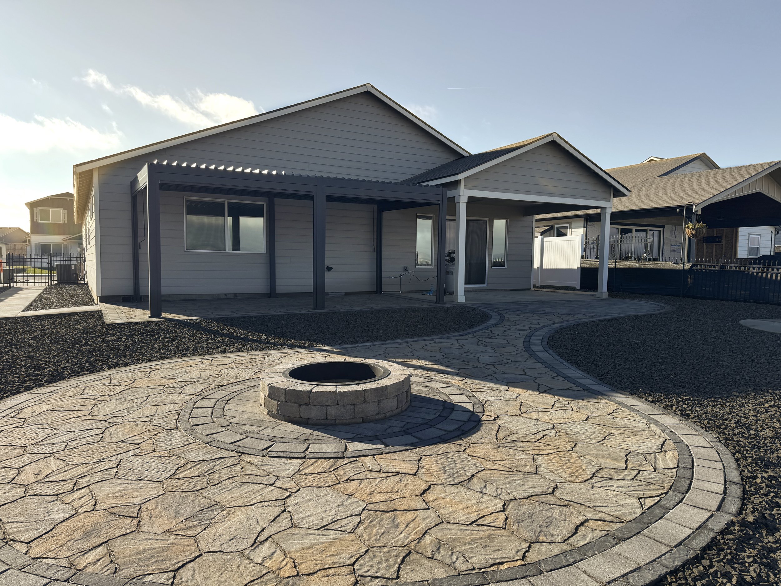 A modern house with a covered patio and a circular stone fire pit in a backyard with a paved walkway and rock landscaping, under a clear blue sky.