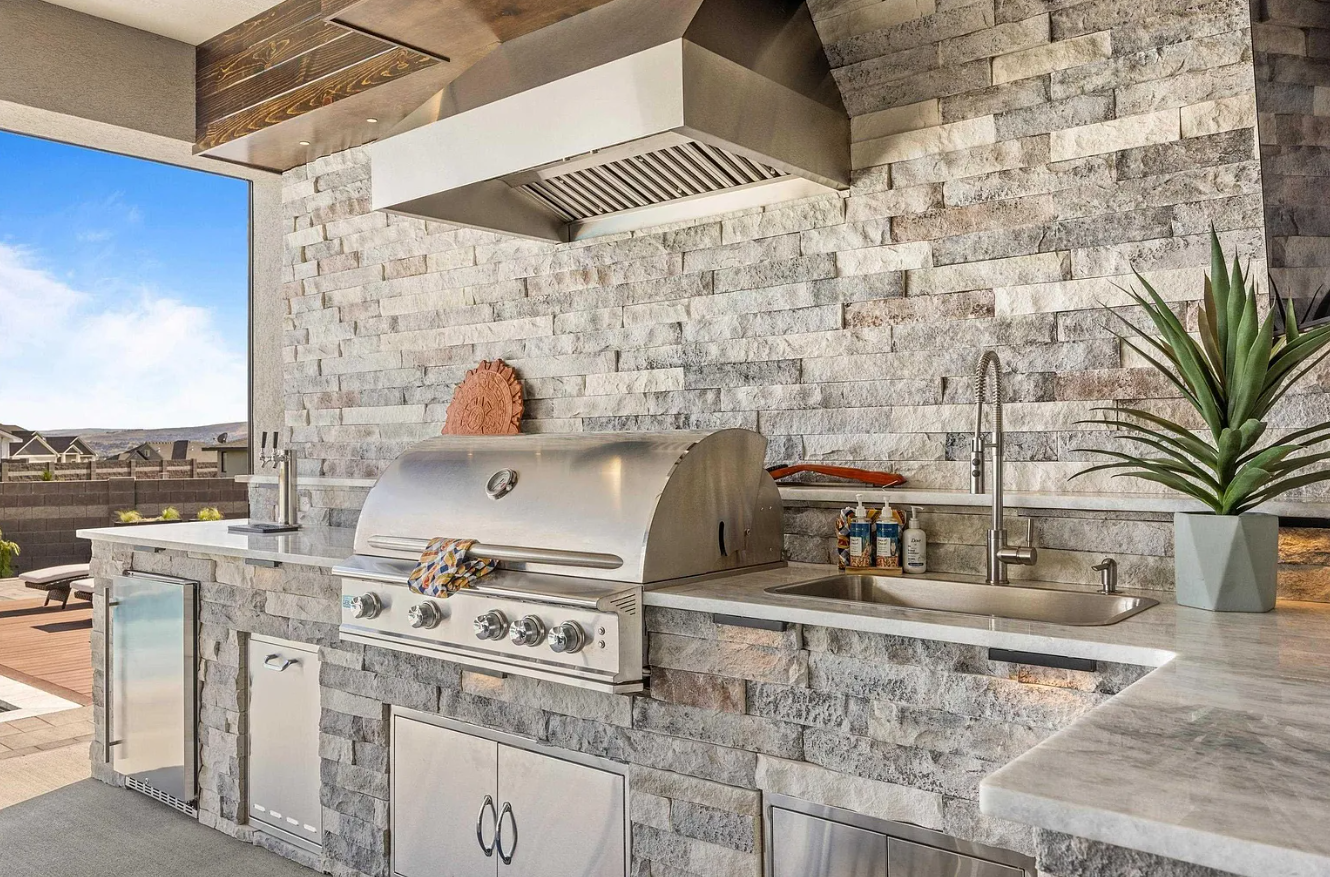 Outdoors kitchen with a stainless steel grill, a sink with a faucet, a potted plant, and a large window showing a backyard with a patio and a blue sky.
