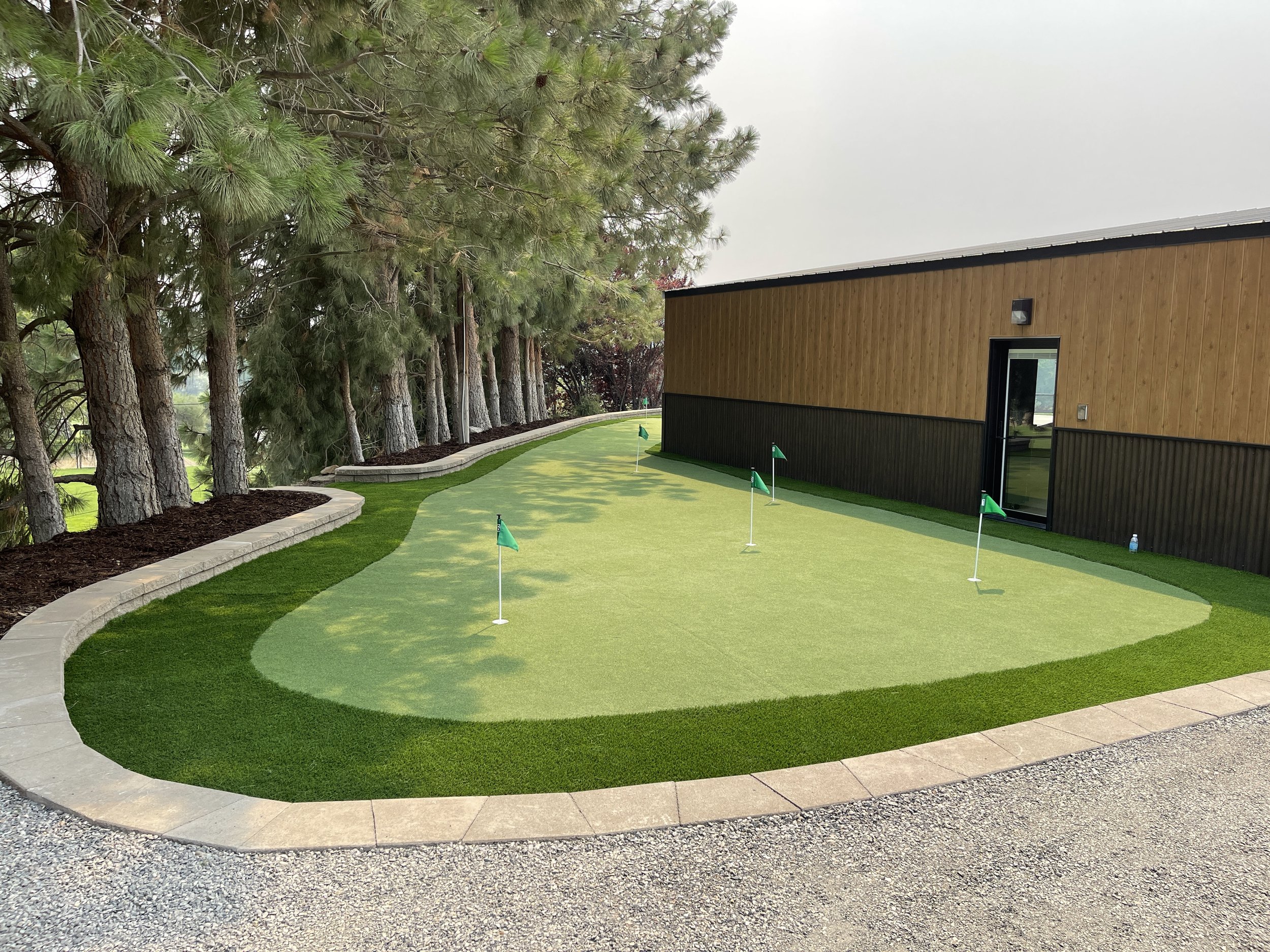 A golf course putting green with several small flags, surrounded by trees and a building with wood siding.