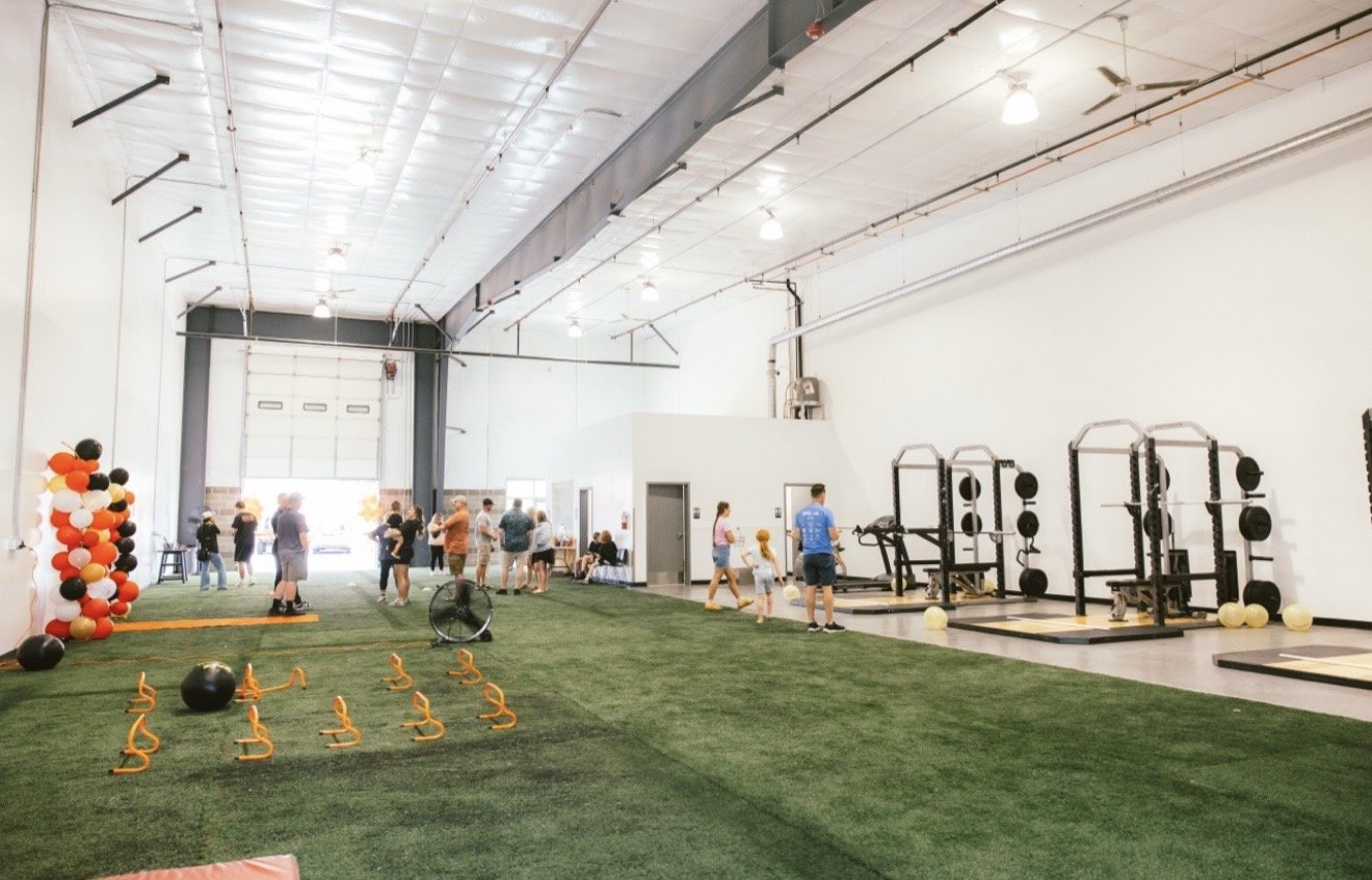 An indoor gym with artificial grass, fitness equipment, and people participating in fitness activities near a large garage door. The space has high ceilings with industrial lighting and decorations with balloons.