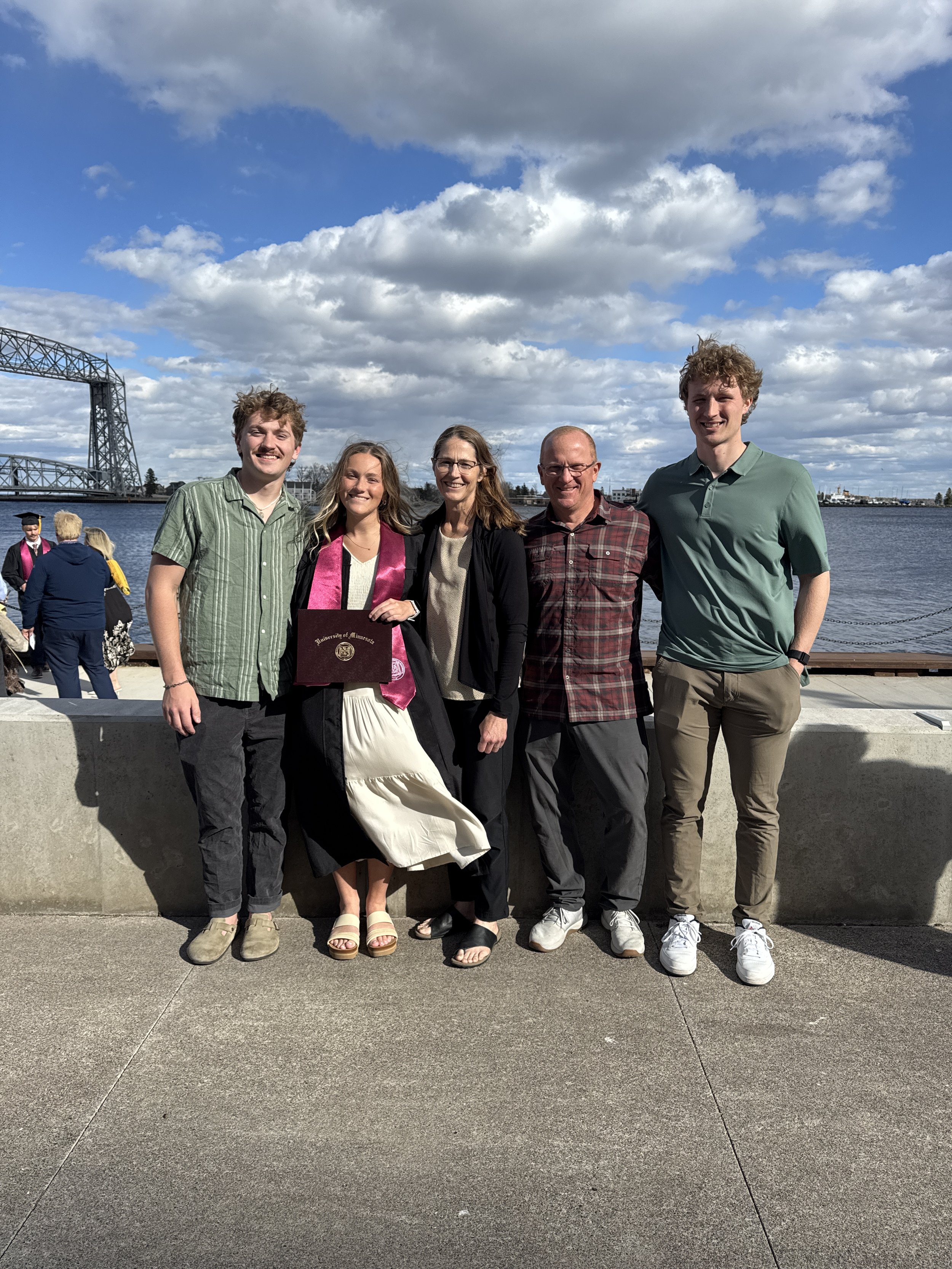 A family group of five standing outdoors by a waterfront, with a bridge and boats in the background. One person is wearing a graduation gown and holding a diploma with a University of Minnesota logo. The group is smiling and dressed casually.