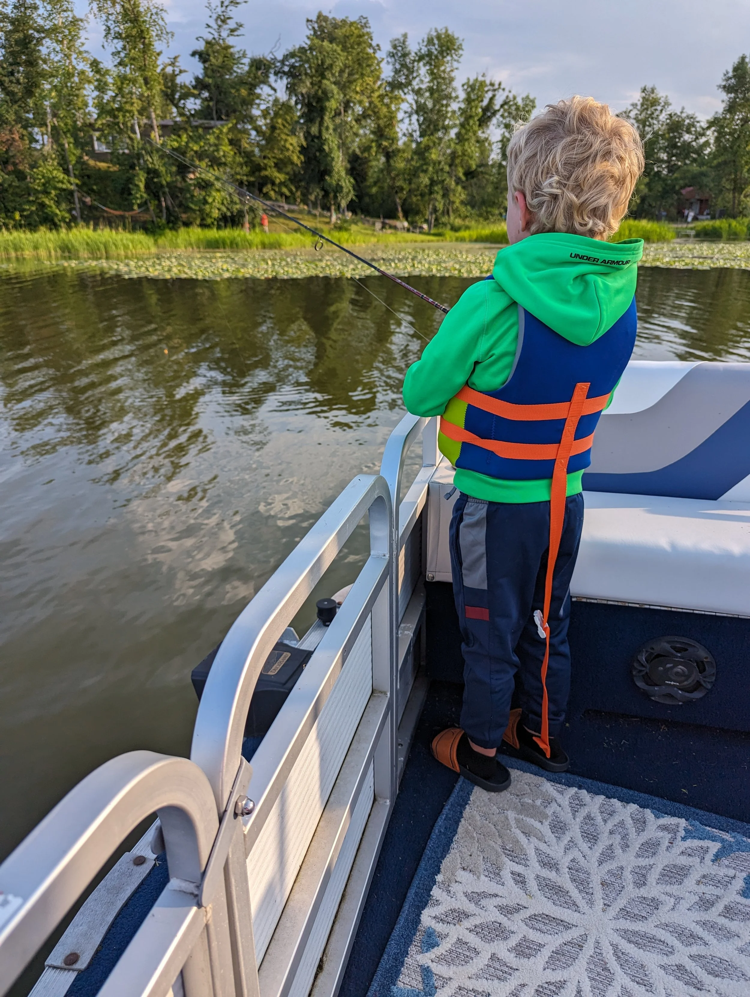 A young boy with blonde hair wearing a bright green and blue life jacket, dark pants, and orange and black sandals, is fishing from a boat on a lake surrounded by green trees and water lilies.
