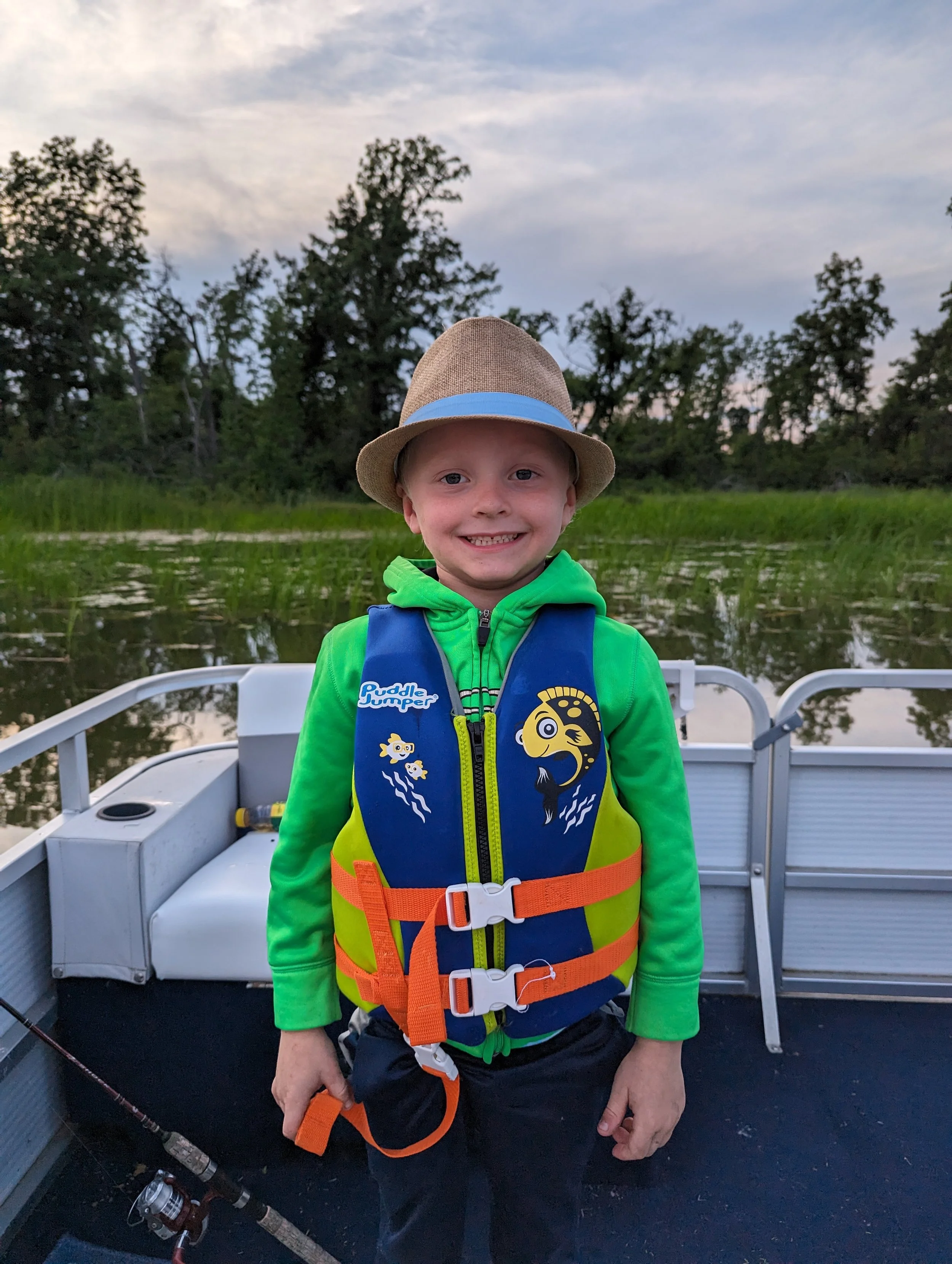 A young boy with a big smile, wearing a straw hat and a fishing life vest, standing on a boat with a fishing rod in his hand, surrounded by water and trees at sunset on White Ash Lake