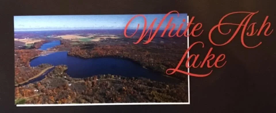 Aerial view of White Ash Lake surrounded by fall foliage with fields in the distance, overlaid with red text 'White Ash Lake'.