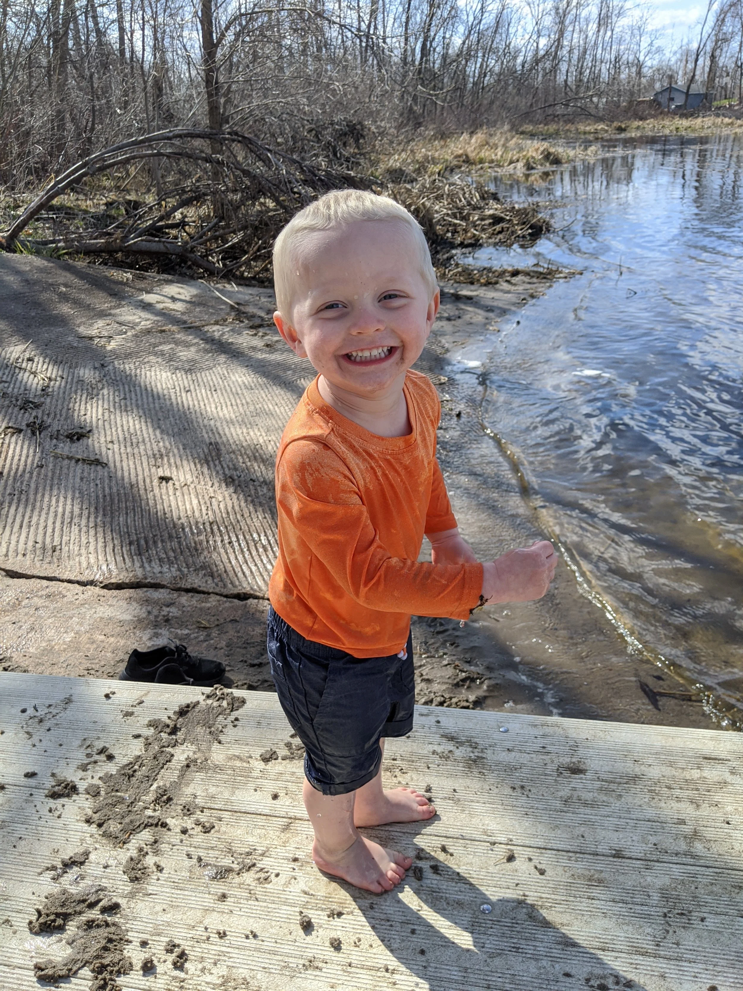 A young boy standing barefoot on a wooden dock by a body of water, smiling widely at the camera during daytime.