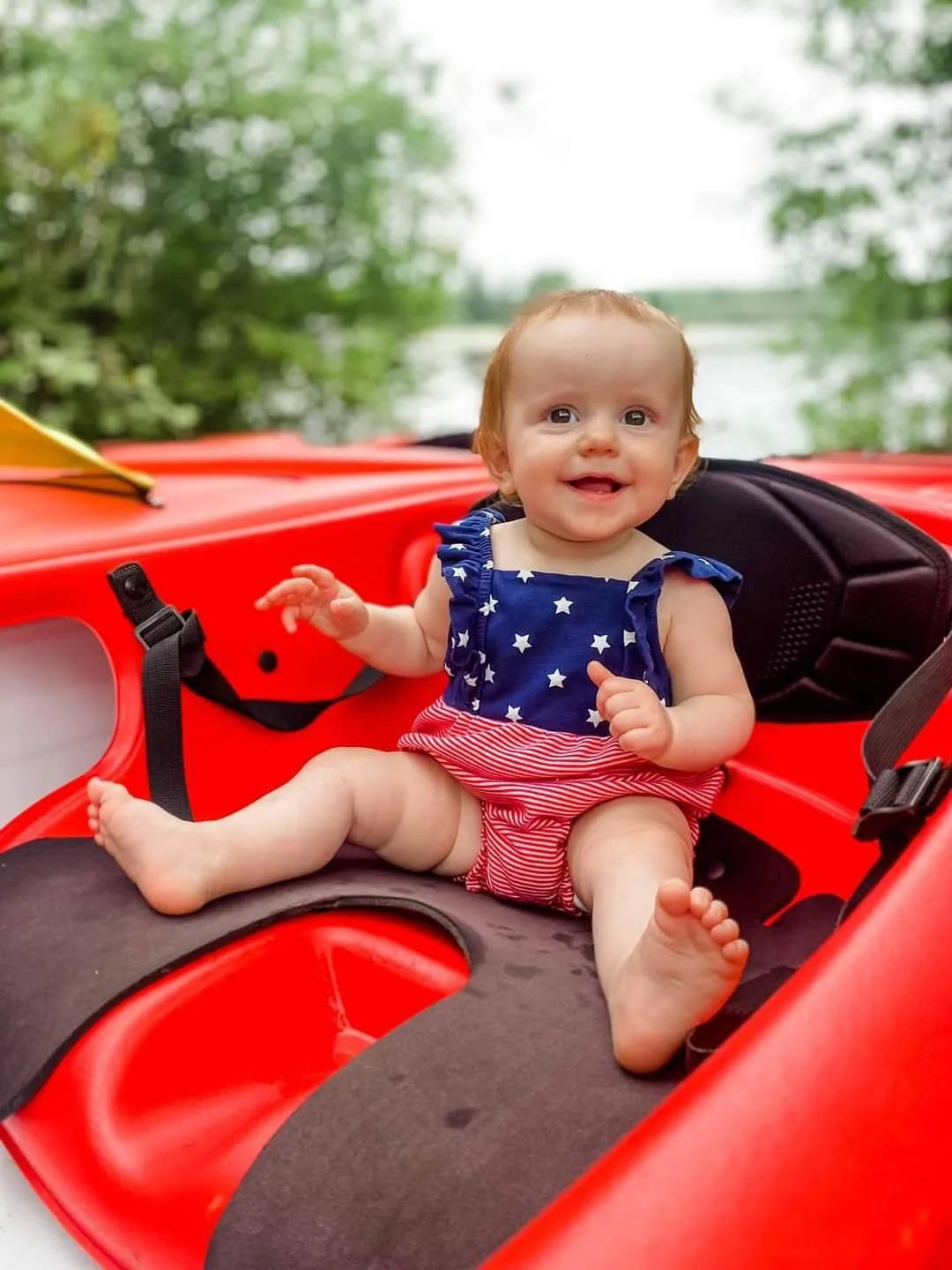 Smiling baby with red hair in patriotic blue and red outfit sitting in a red kayak outdoors by a lake with trees and water in the background.