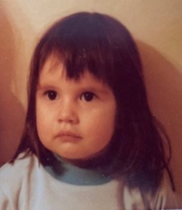 A young girl with dark hair and bangs, wearing a light-colored shirt, looking slightly to the side, against a neutral background.