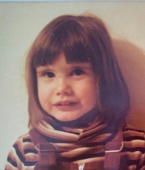 A young girl with straight, shoulder-length brown hair and a striped shirt, looking at the camera.