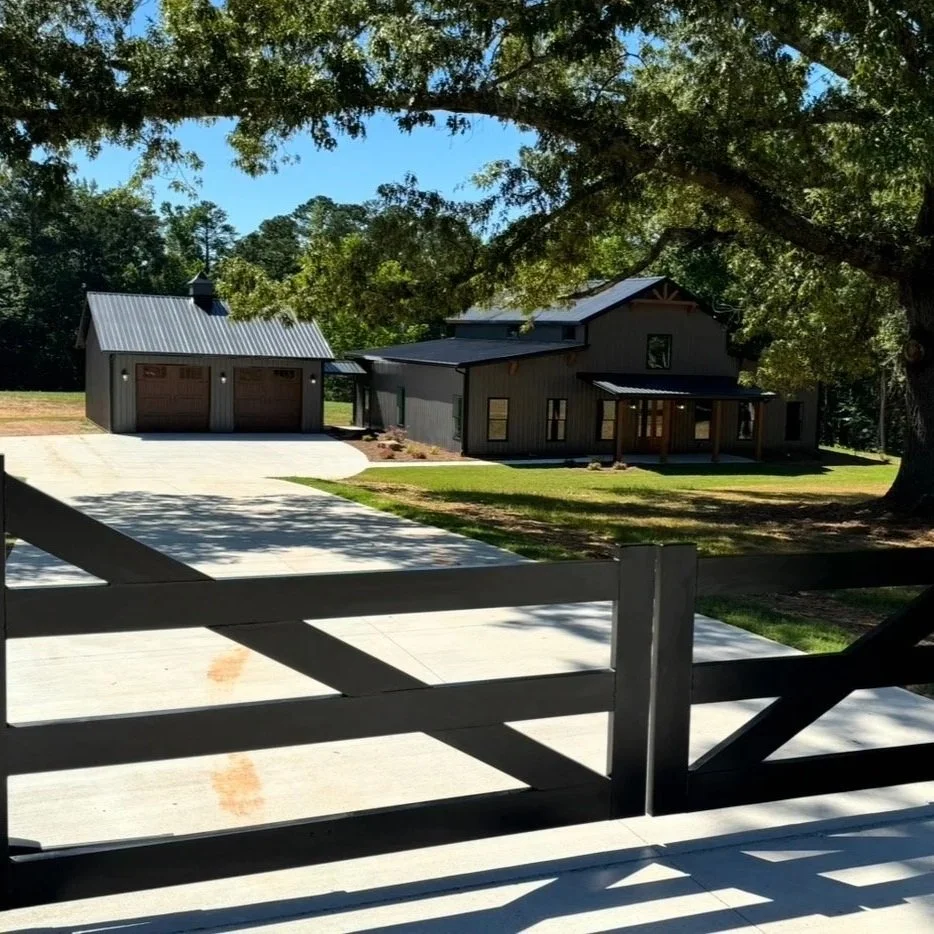 A modern house with a metal roof, a detached garage with two doors, surrounded by trees and a well-maintained lawn, seen from a porch with a wooden fence.