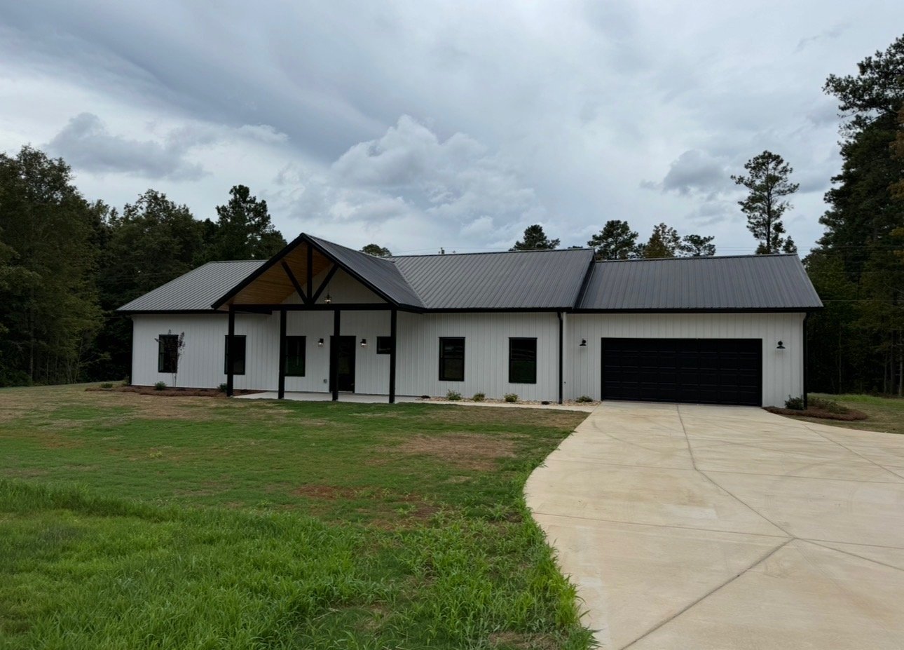 Modern farmhouse-style house with white exterior, black trim, metal roof, attached garage, and a curved concrete driveway surrounded by green grass and trees under a cloudy sky.