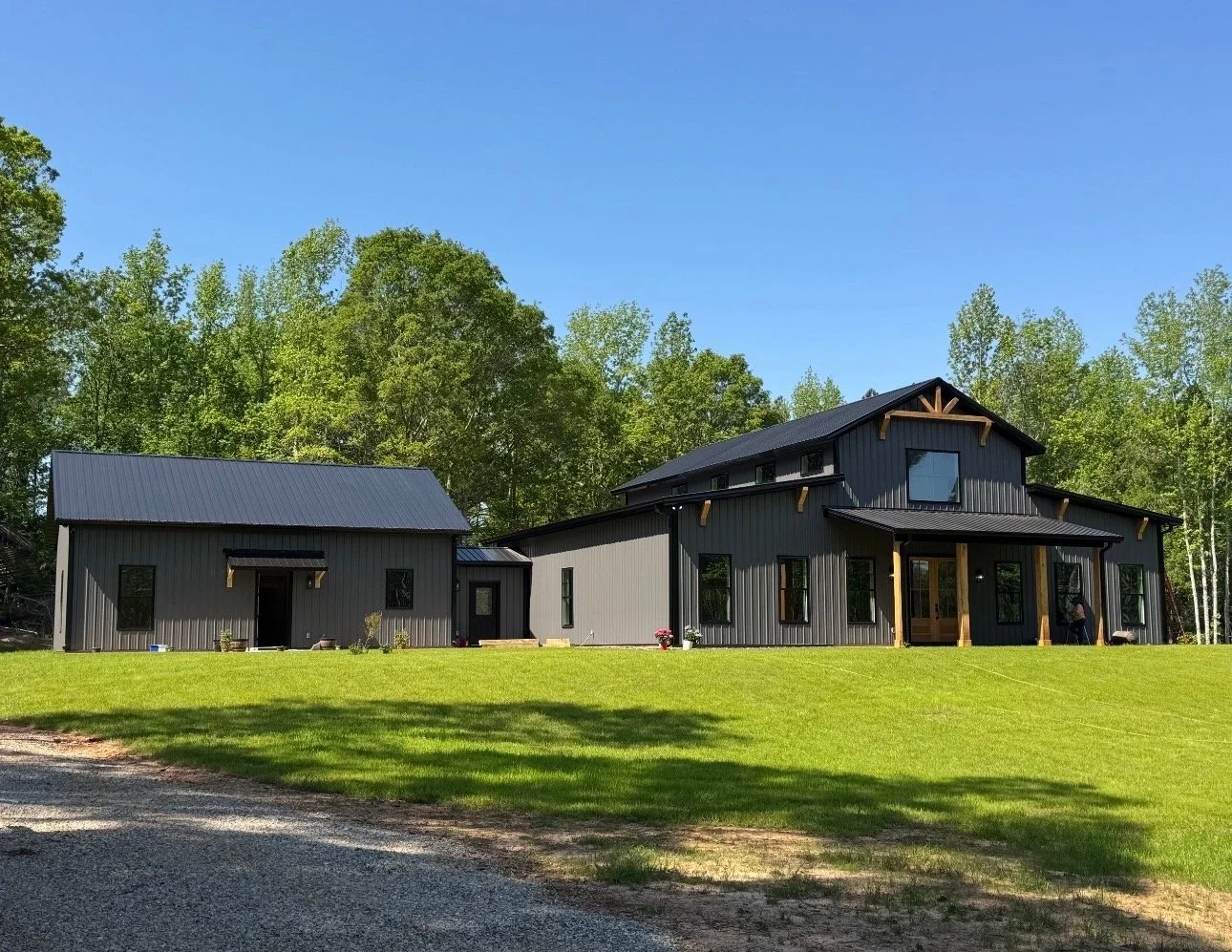 Modern black house with metal siding, large windows, and a front porch, set on a grassy yard with trees in the background and a gravel driveway in the foreground.
