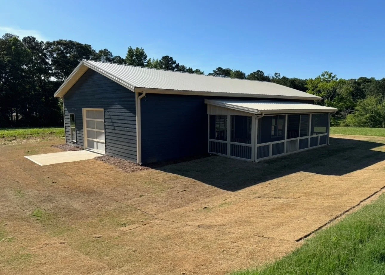 Newly constructed blue house with a screened porch and metal roof, surrounded by a dry yard and green trees in the background.