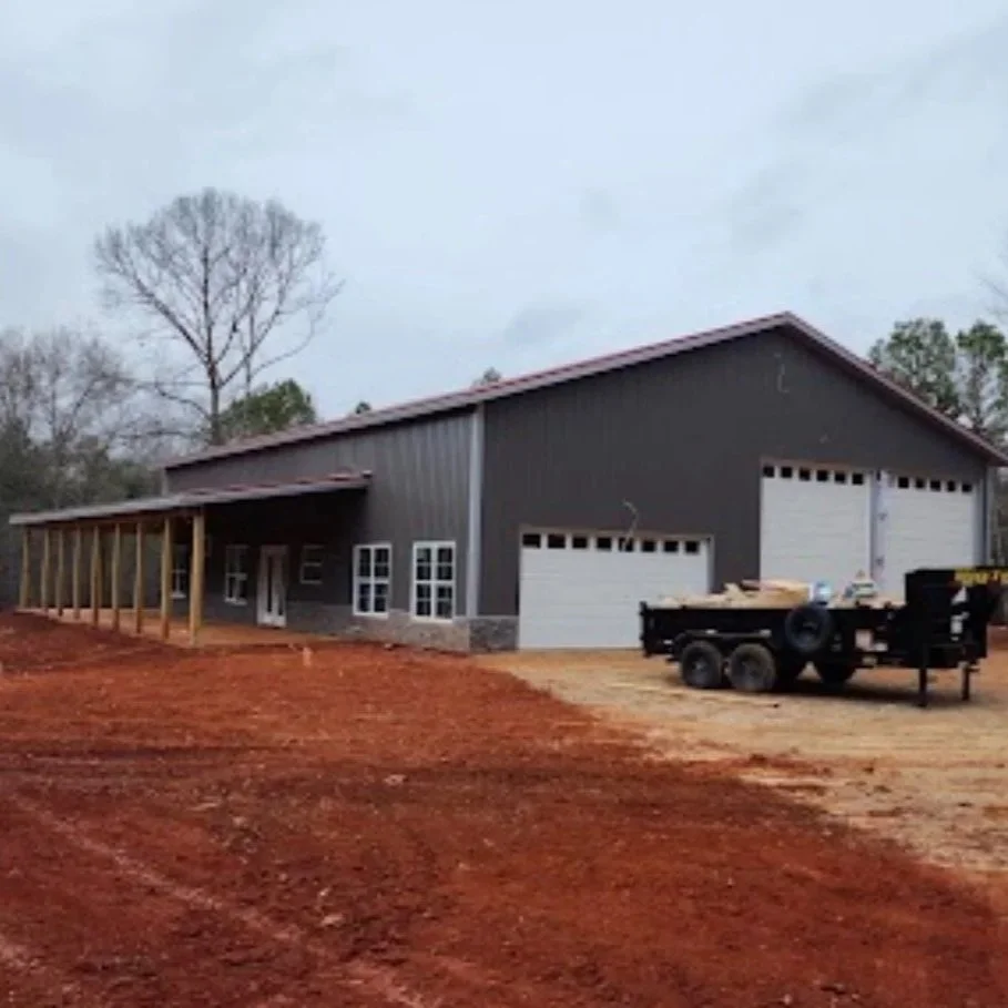 A large dark gray metal barn with two garage doors, a small porch area with white double doors, and an attached covered area under construction. The ground is reddish dirt, and there is a trailer with supplies parked in front. Bare trees are in the background.