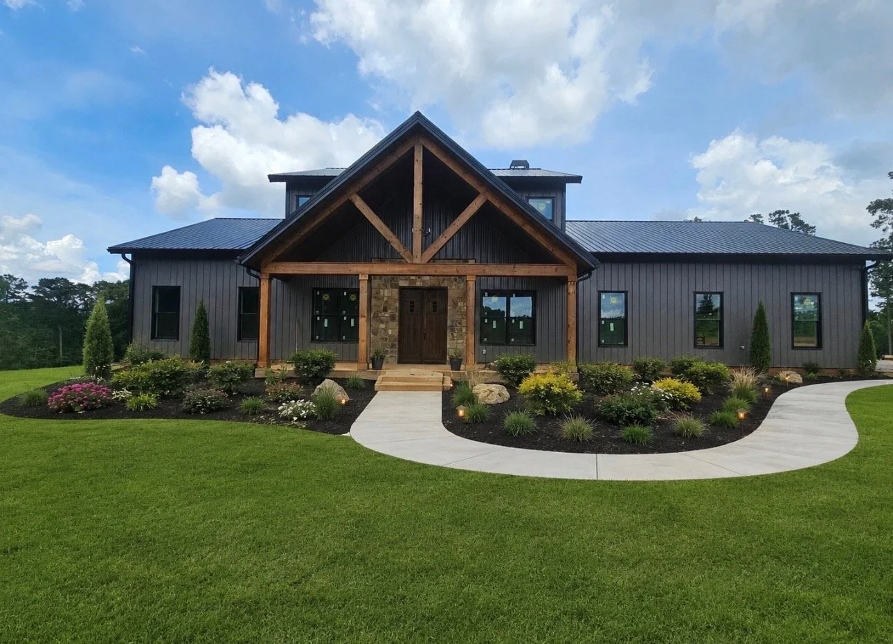 A modern farmhouse-style house with gray siding, wooden beams, and stone accents, surrounded by a well-maintained lawn and landscaped garden with shrubs and flowers.