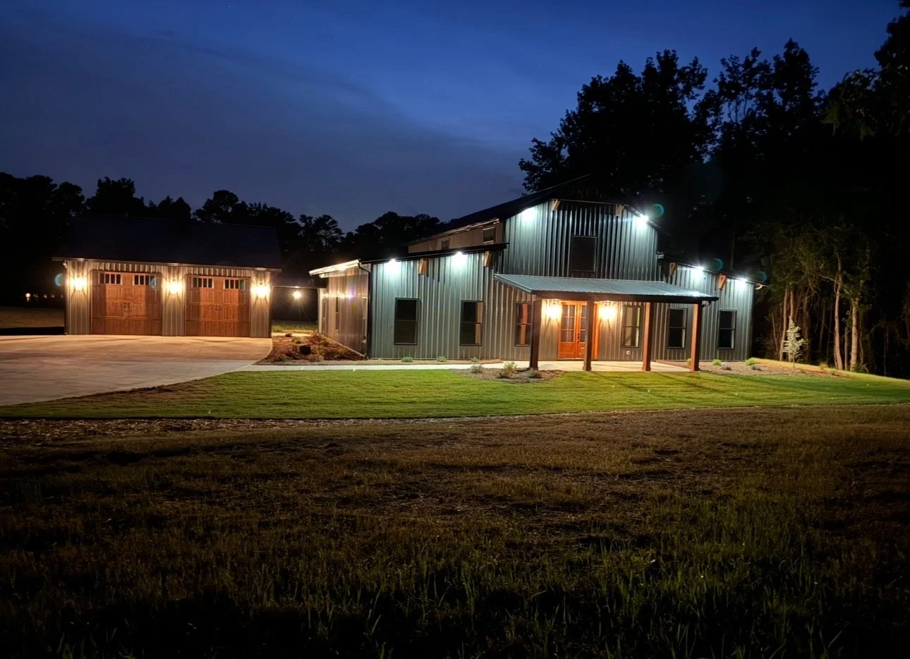 A large metal barn-like house illuminated at night, with a separate garage nearby, surrounded by a grassy yard and trees in the background.