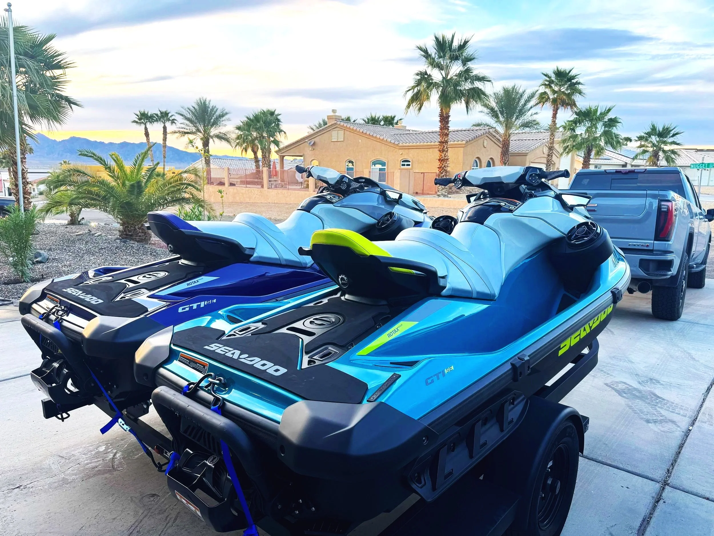 Two Yamaha jet skis with blue, black, white, and neon yellow accents parked on a driveway in front of a desert-style house with palm trees and mountains in the background.