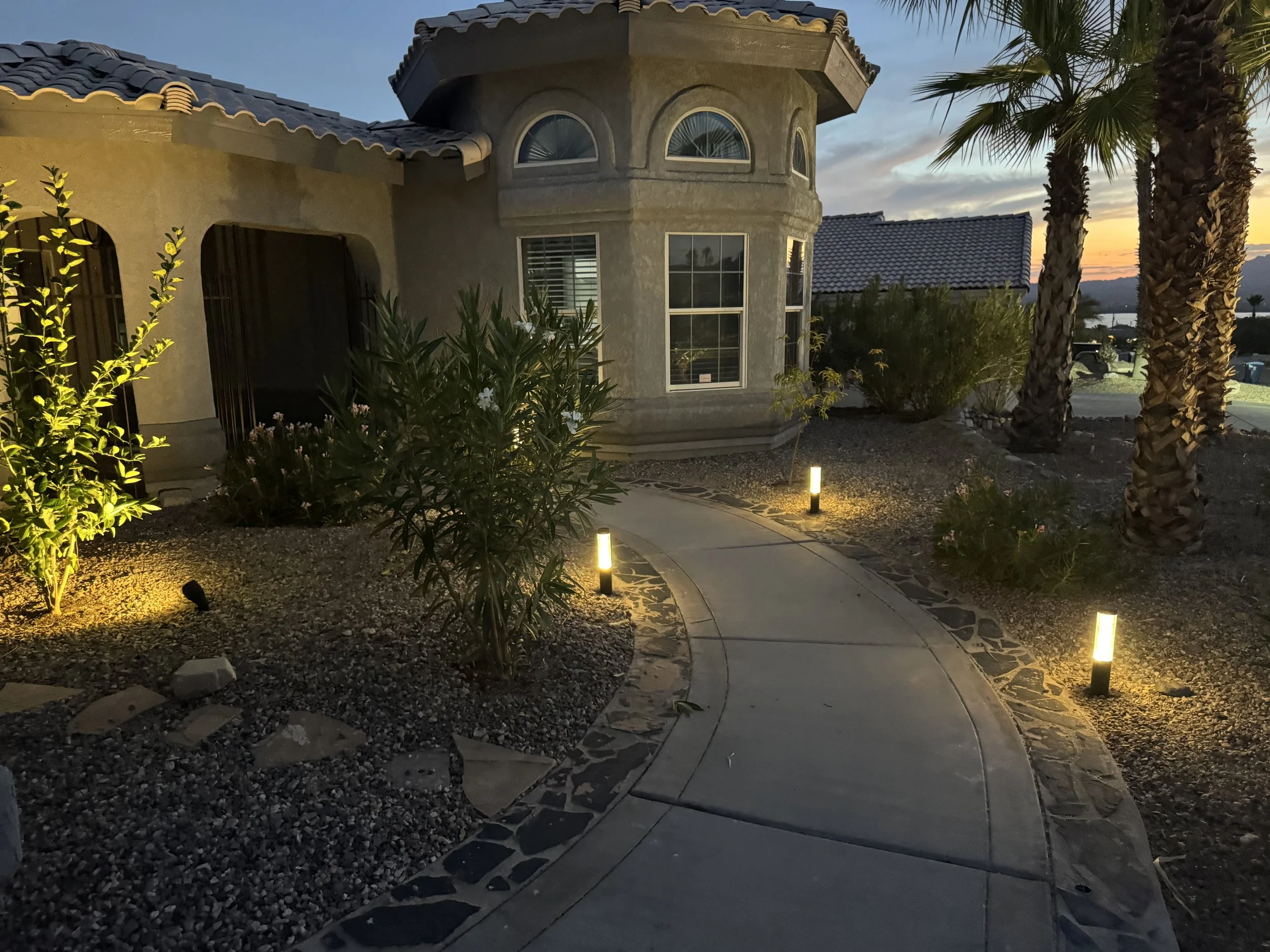 A house with a textured exterior and arched windows at sunset, surrounded by palm trees and landscaped yard illuminated with pathway lights.