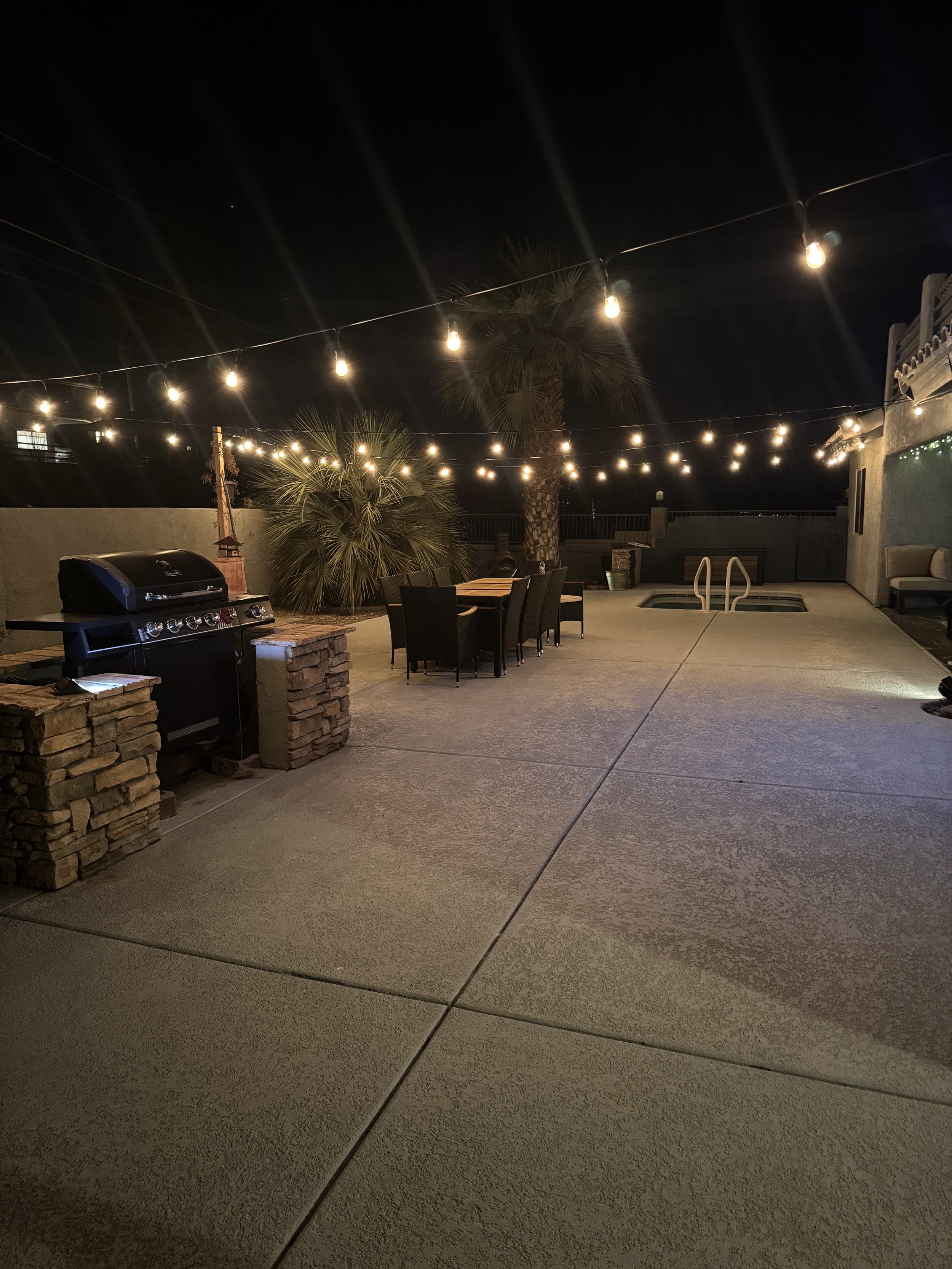 Nighttime backyard patio scene with string lights hanging above, a table with black chairs, a barbecue grill, and palm trees in the background.