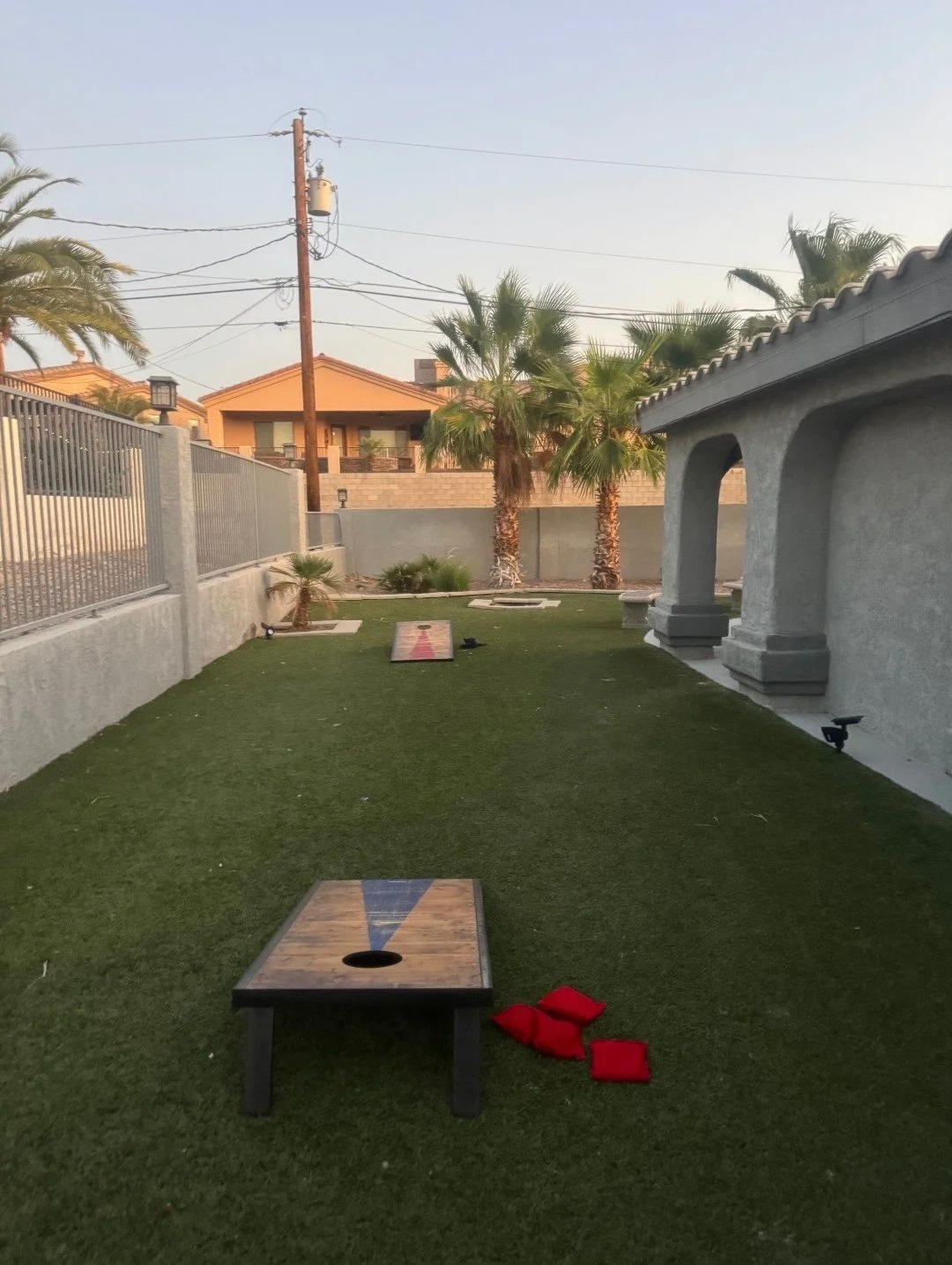 Backyard with artificial grass, cornhole game, palm trees, and a house with stucco walls and arches.