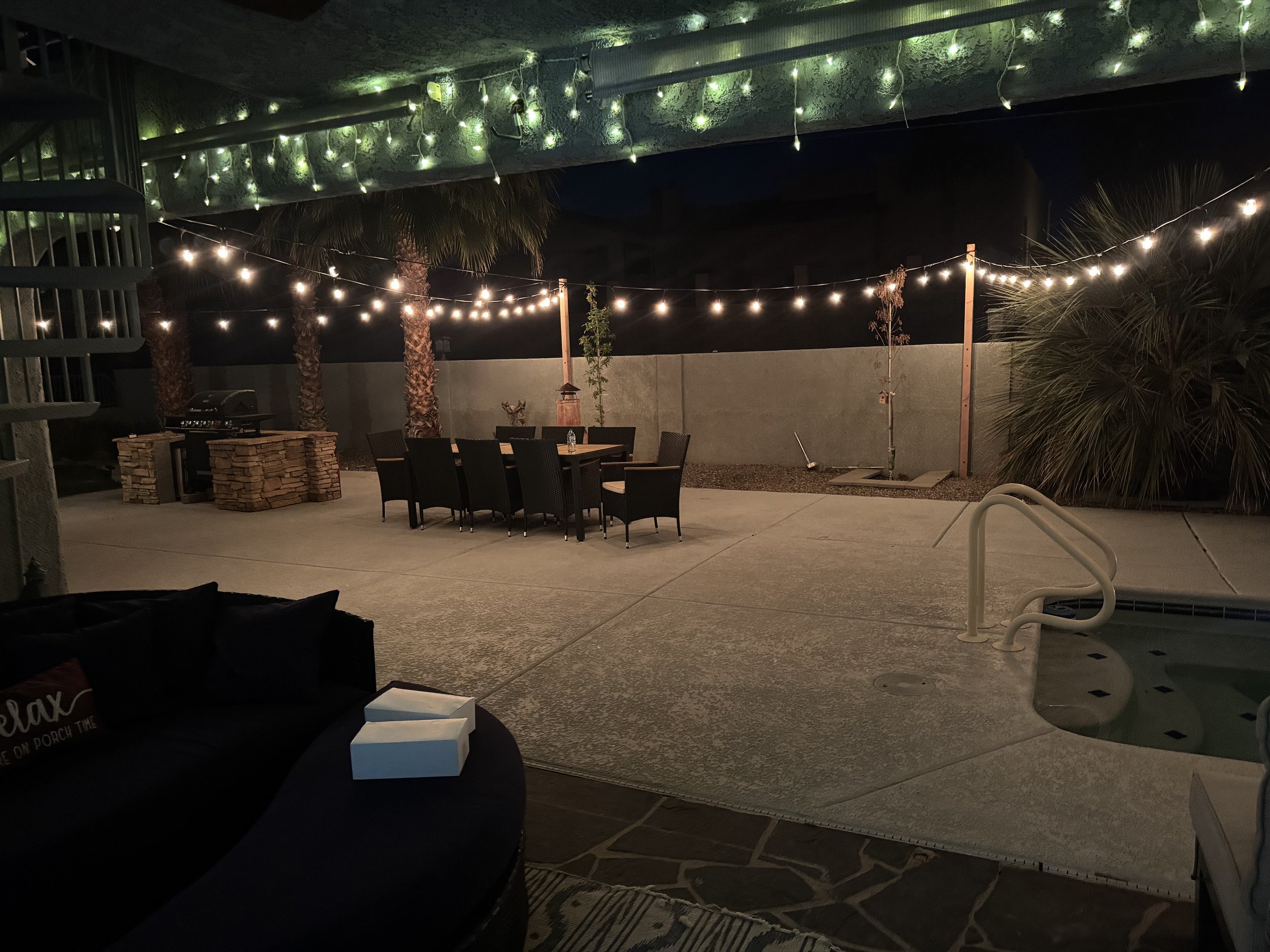 An outdoor patio at night with string lights hanging overhead. There are palm trees, a table with chairs, and a stone firepit. The area is lit with small green and white lights, and there's a pool edge visible in the foreground.