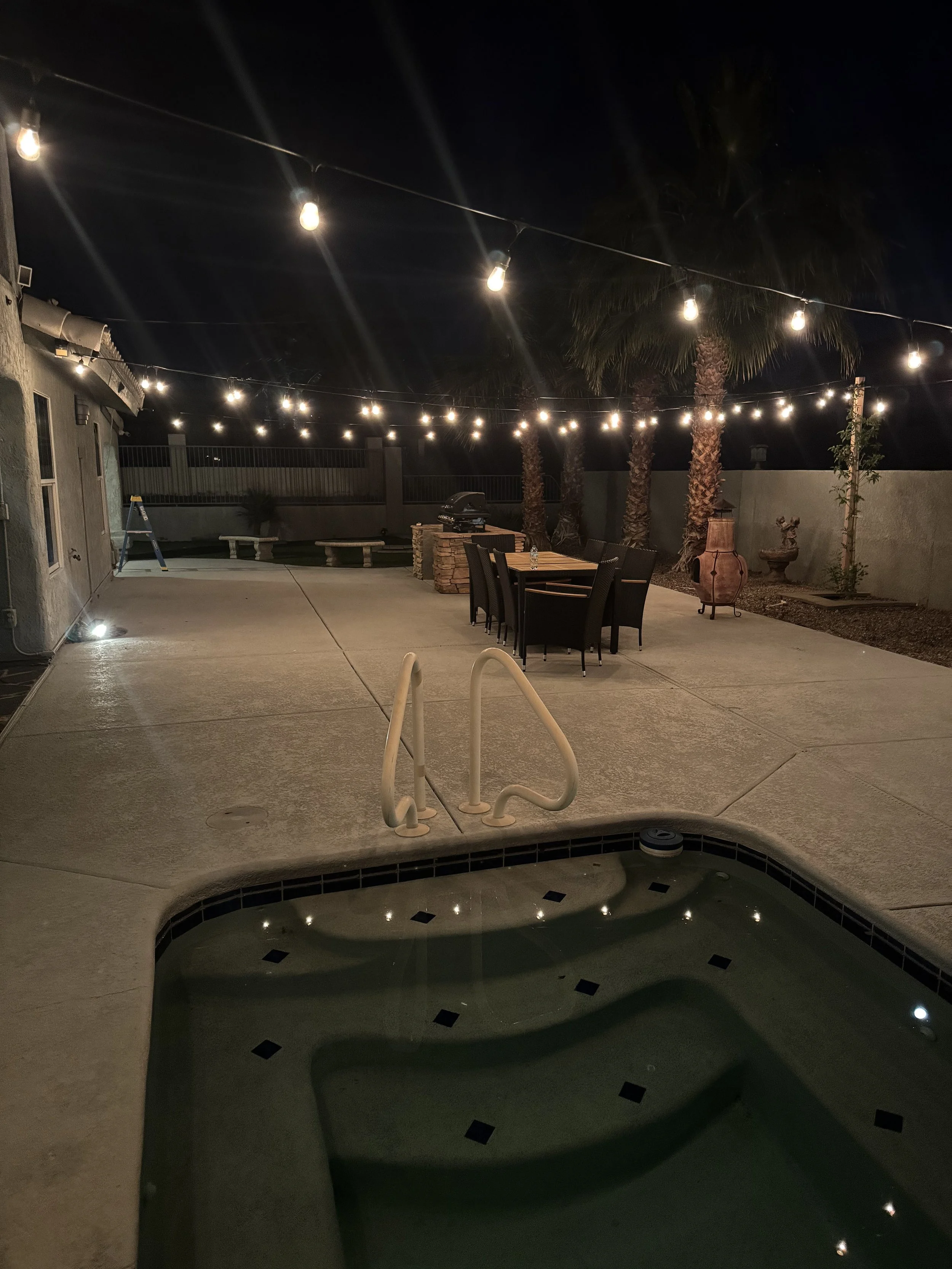 Night view of a backyard patio with string lights overhead, a small hot tub in the foreground, outdoor seating, a barbecue grill, and palm trees.