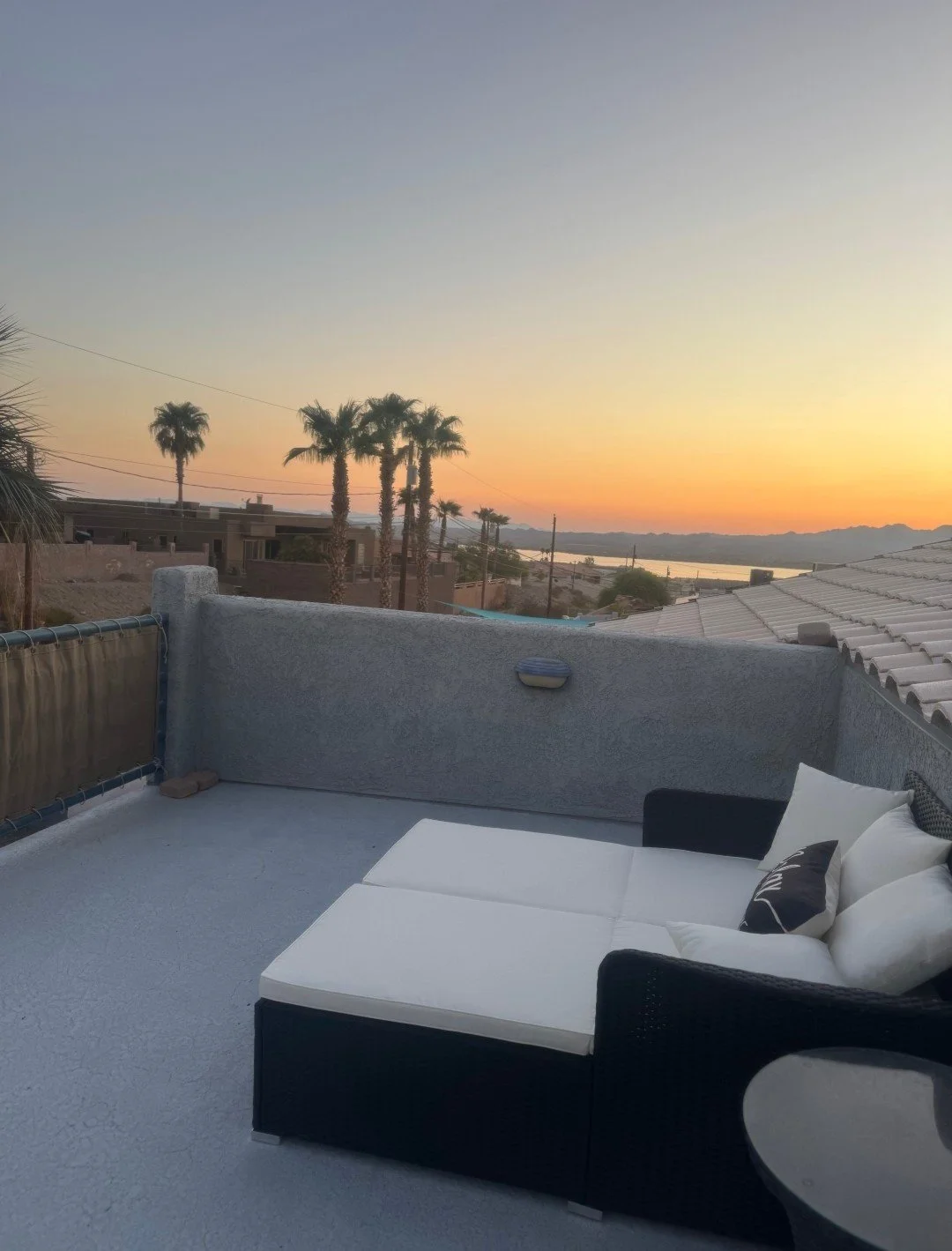 A rooftop patio with a black and white lounge chair and pillows, overlooking a desert landscape with palm trees and a body of water at sunset.