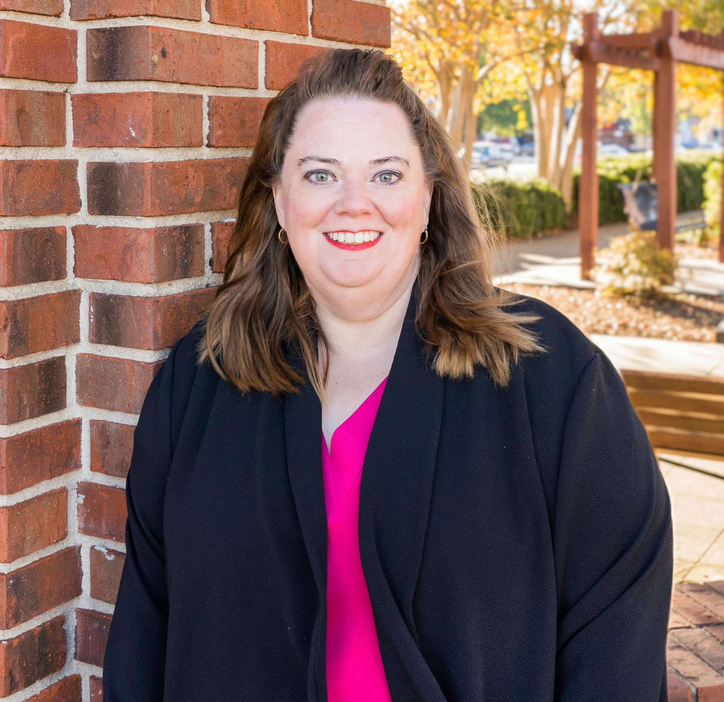 A woman with long, wavy brown hair and green eyes smiling, wearing a black jacket and a bright pink shirt underneath, standing outdoors against a brick wall with autumn trees and a park bench in the background.