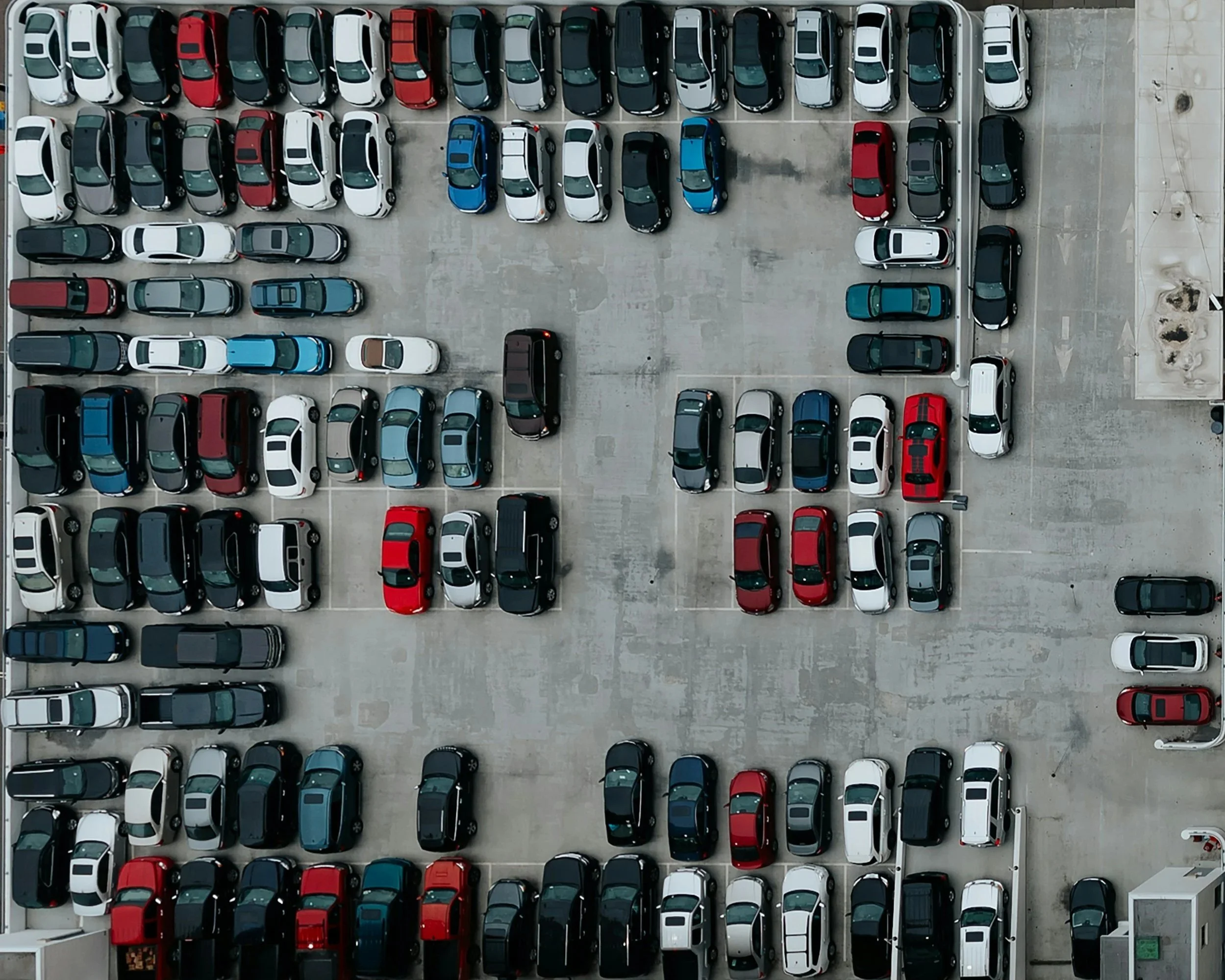 Aerial view of a crowded parking lot with multiple rows of parked cars, including various colors like white, black, red, blue, and gray.