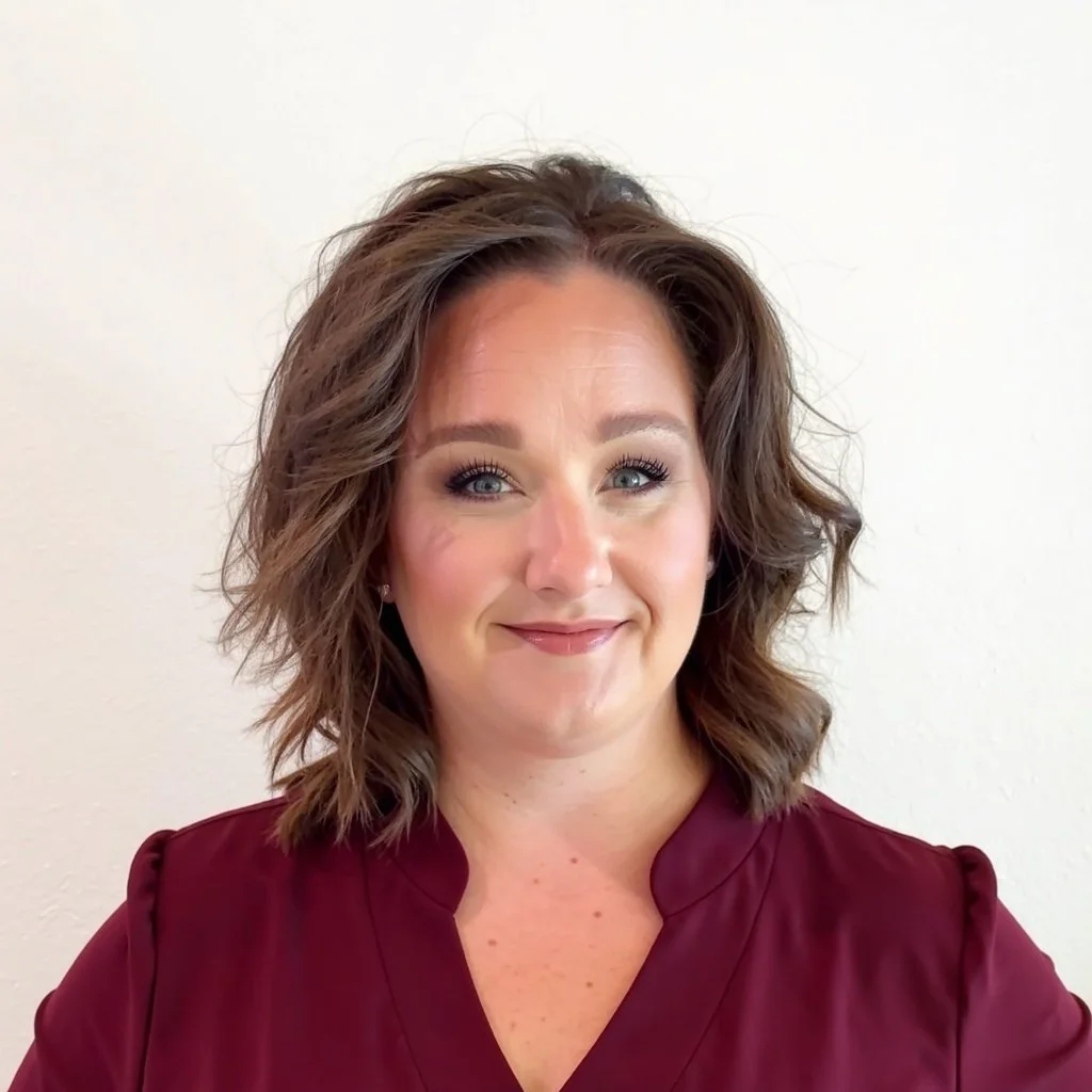 A woman with shoulder-length brown hair and blue eyes, smiling, wearing a maroon blouse, standing against a plain white wall.