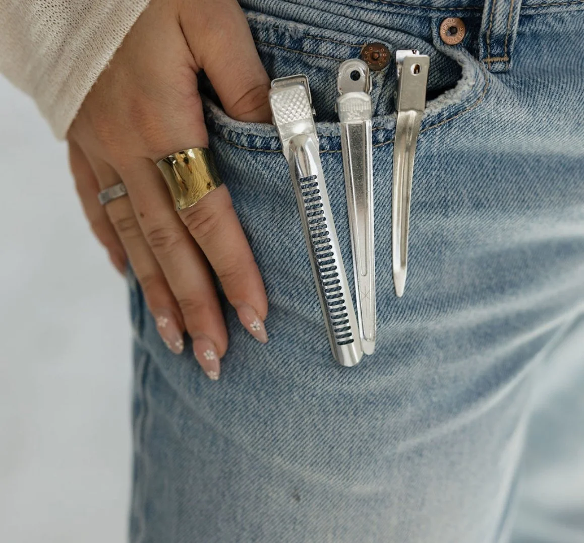 Close-up of a person in blue jeans with five hairdressing tools in the front pocket, including a tweezers, a nail file, a cuticle pusher, a small box cutter, and another object, with a hand resting on the hip and wearing rings and a gold bracelet.