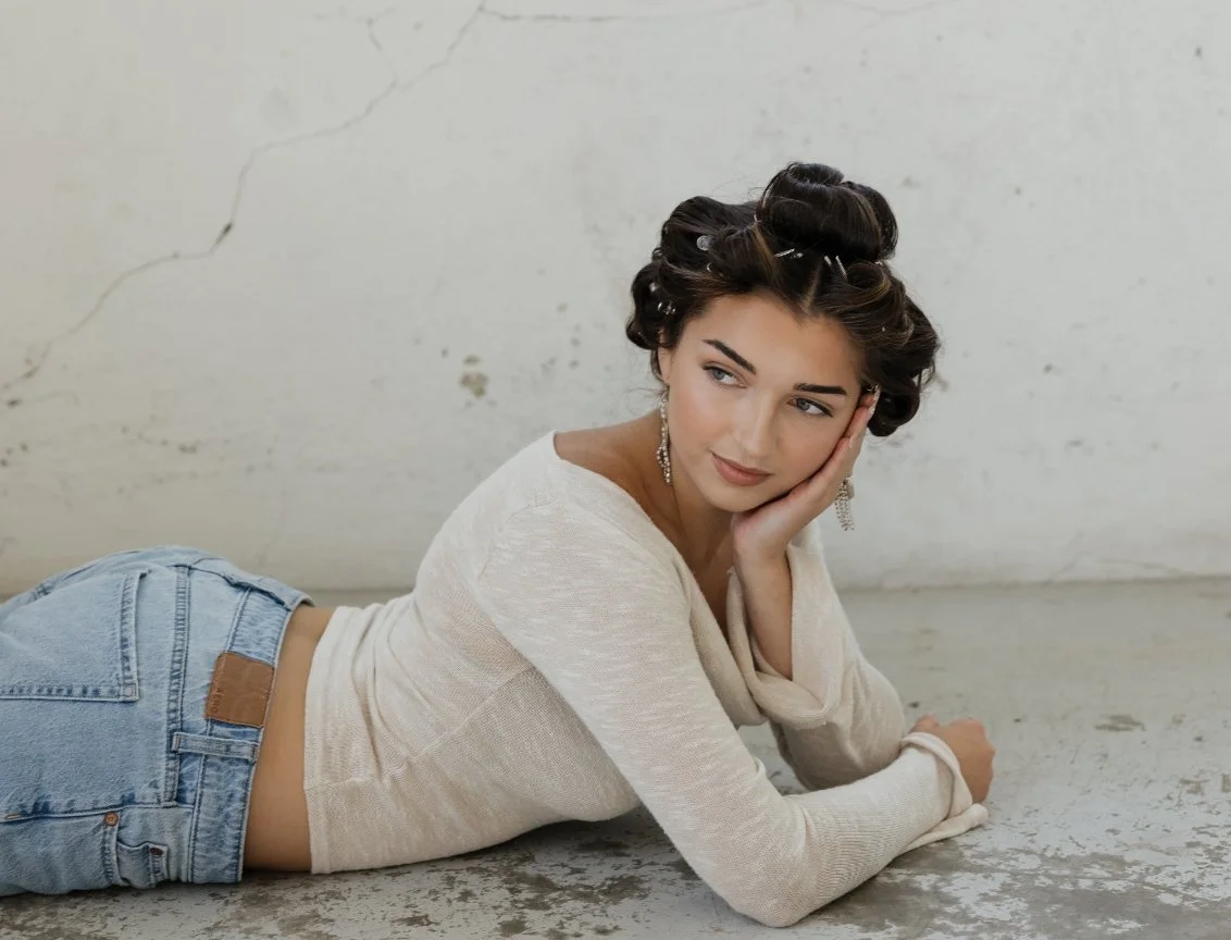A young woman with dark hair pinned up, wearing a beige top and blue jeans, lying on her side on a concrete floor with a white wall background, resting her head on her hand and looking sideways.