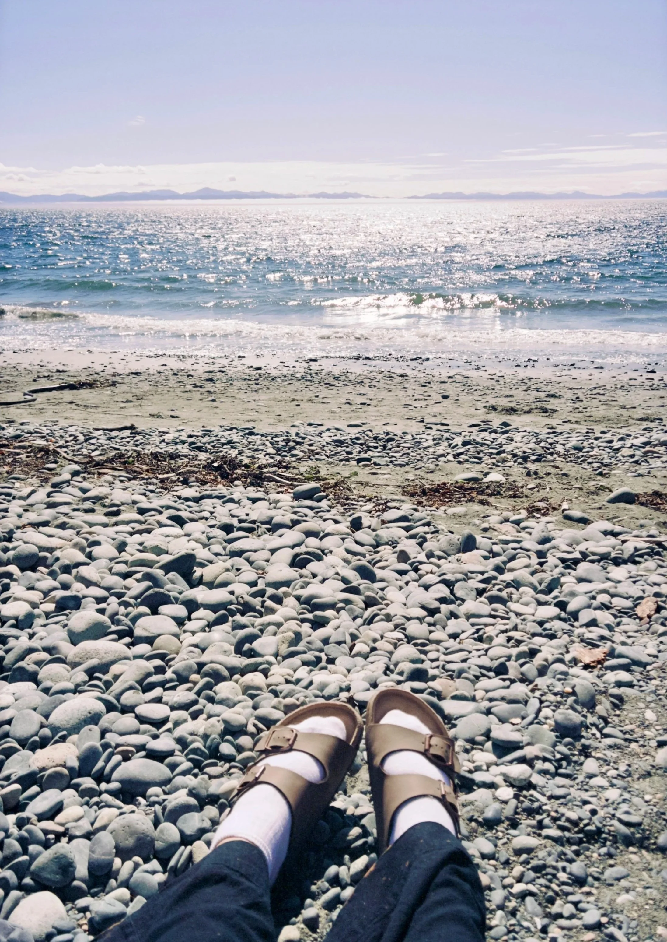 A person in sandals and socks sitting on a pebble-covered beach, looking out at the ocean with waves and distant mountains.